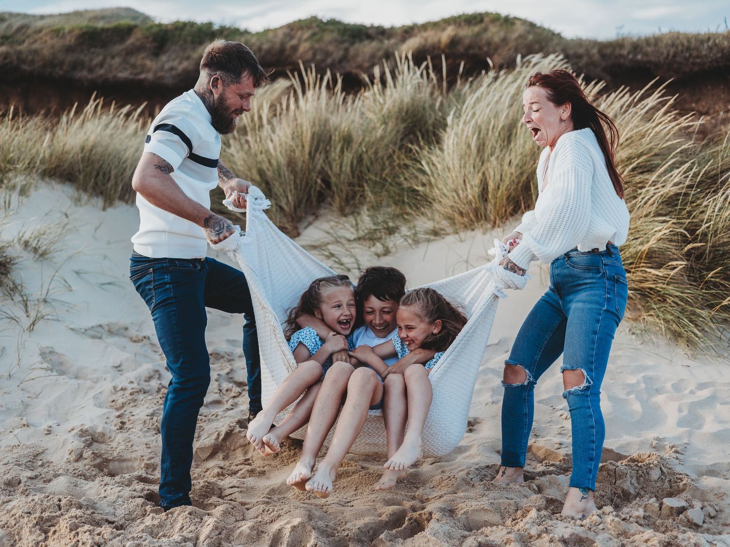 Family of five playing on sandy beach dunes, parents swinging children in a white blanket, laughing outdoors.