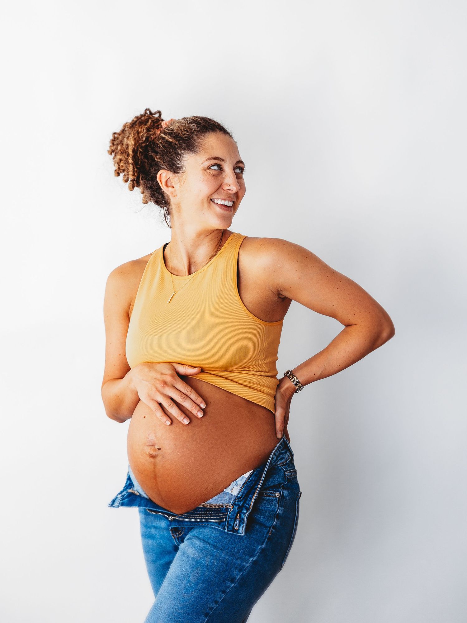 A pregnant woman in a yellow tank top and jeans laughs joyfully while touching her baby bump against a white background.