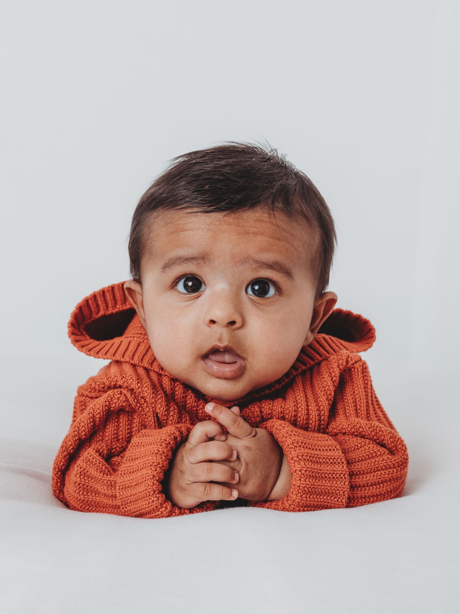 A baby peeks playfully over an orange knit sweater in a series of four candid studio portraits.
