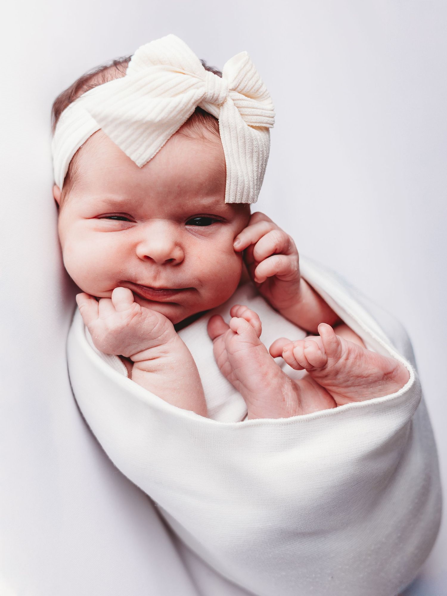 A tiny newborn wrapped in a white blanket wearing a cream-colored bow headband while resting on a light background.