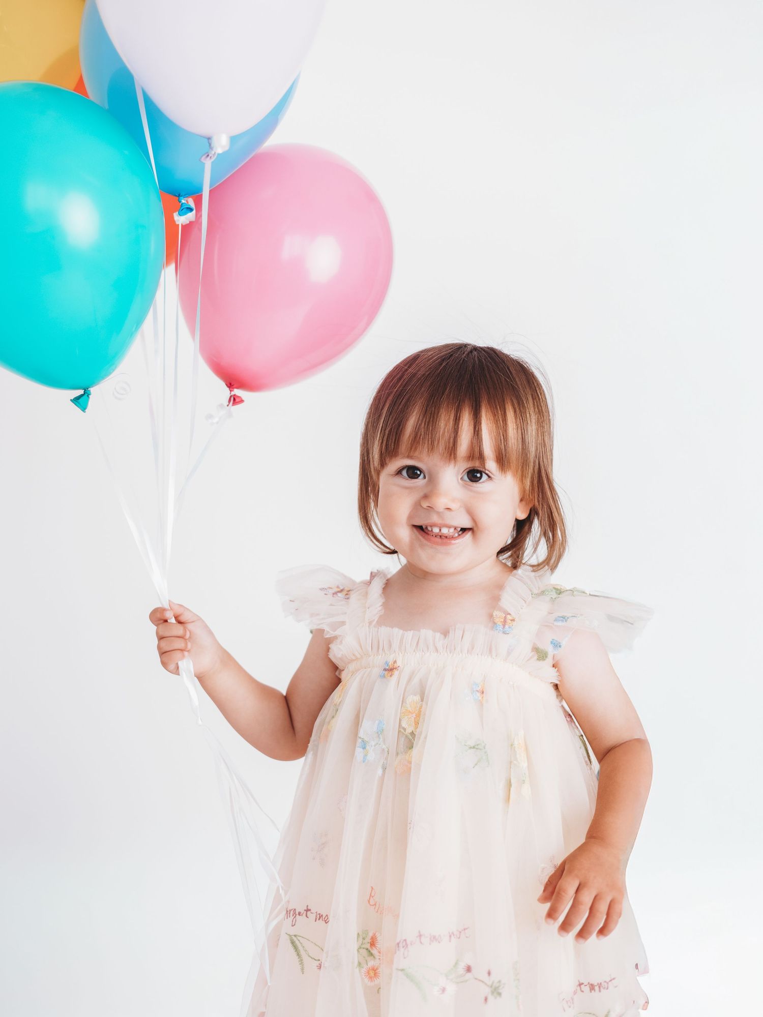 Smiling toddler girl in floral dress holding colorful balloons against white background.
