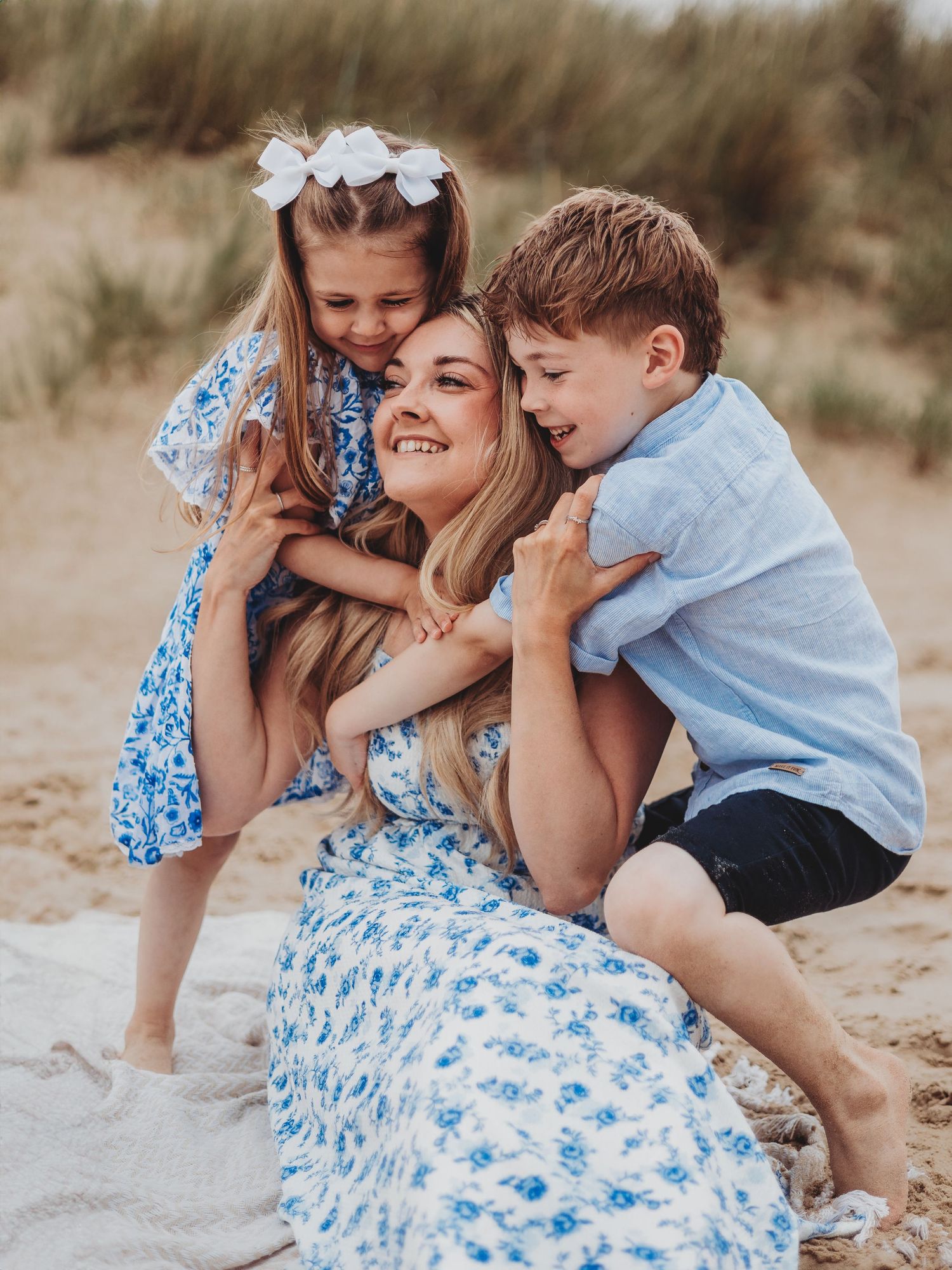 A mother sits on sandy beach smiling as two young children hug her, all wearing blue and white outfits.