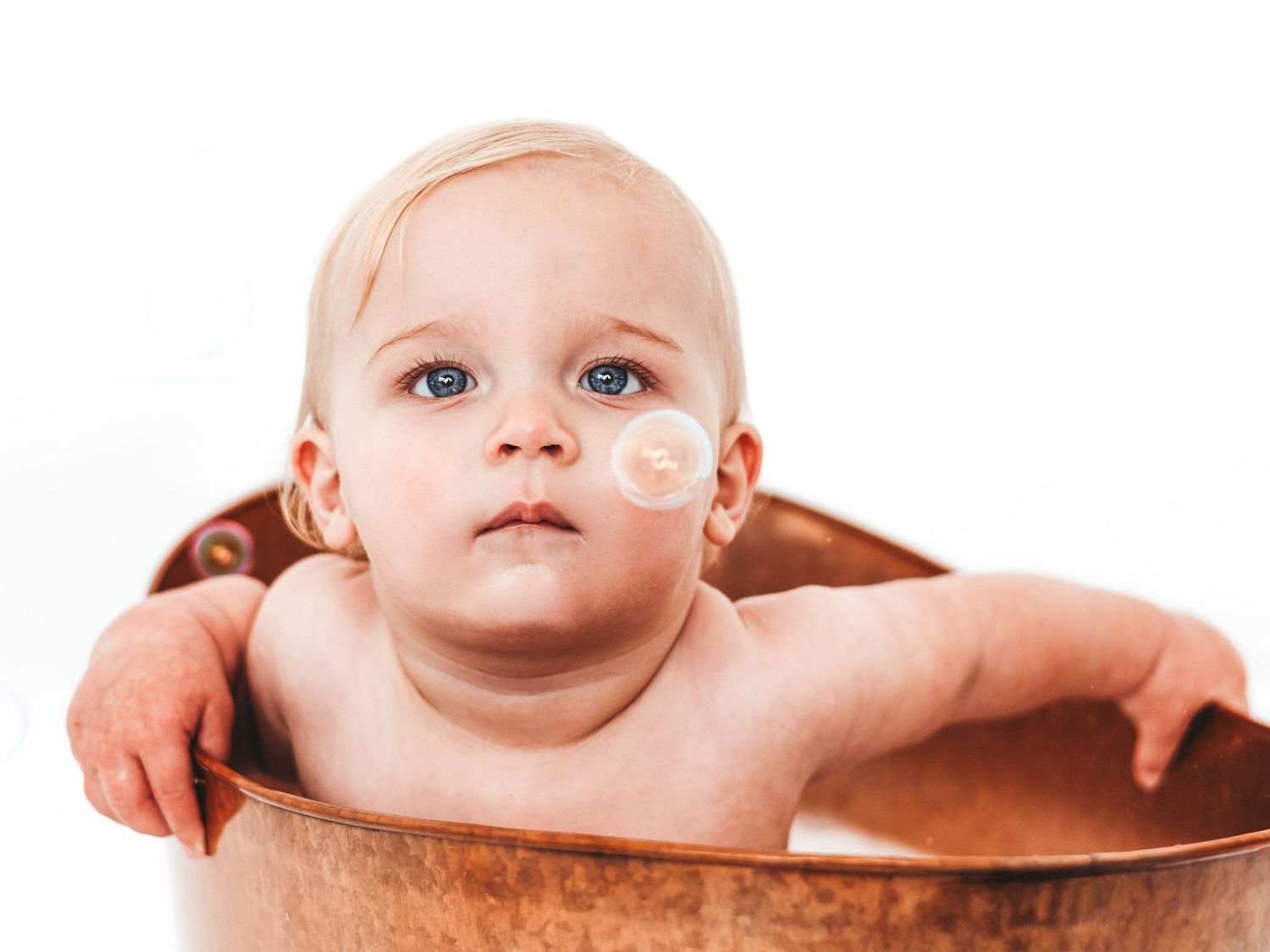 Blue-eyed baby with blonde hair sitting in a copper bathtub against a white background.