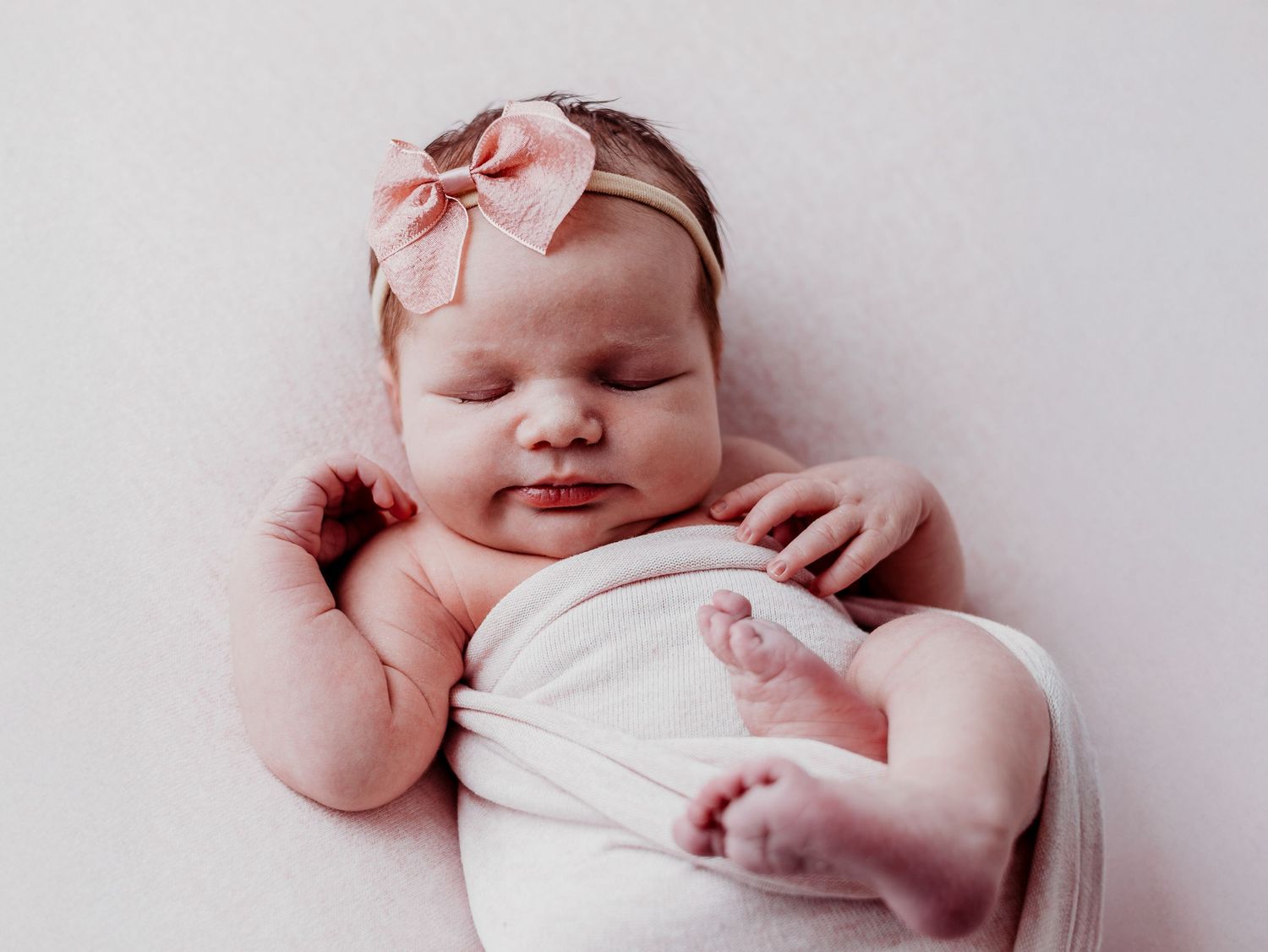 A newborn baby wearing a pink bow headband sleeps peacefully in soft white bedding.