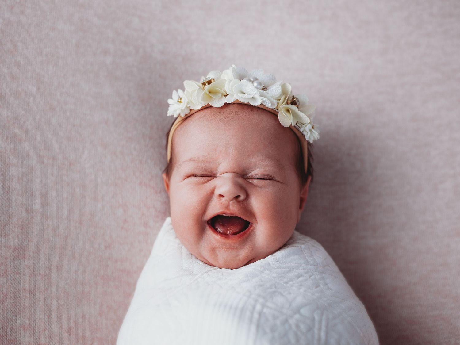 Newborn baby wearing white floral crown smiles while lying on soft gray background during professional photo session.