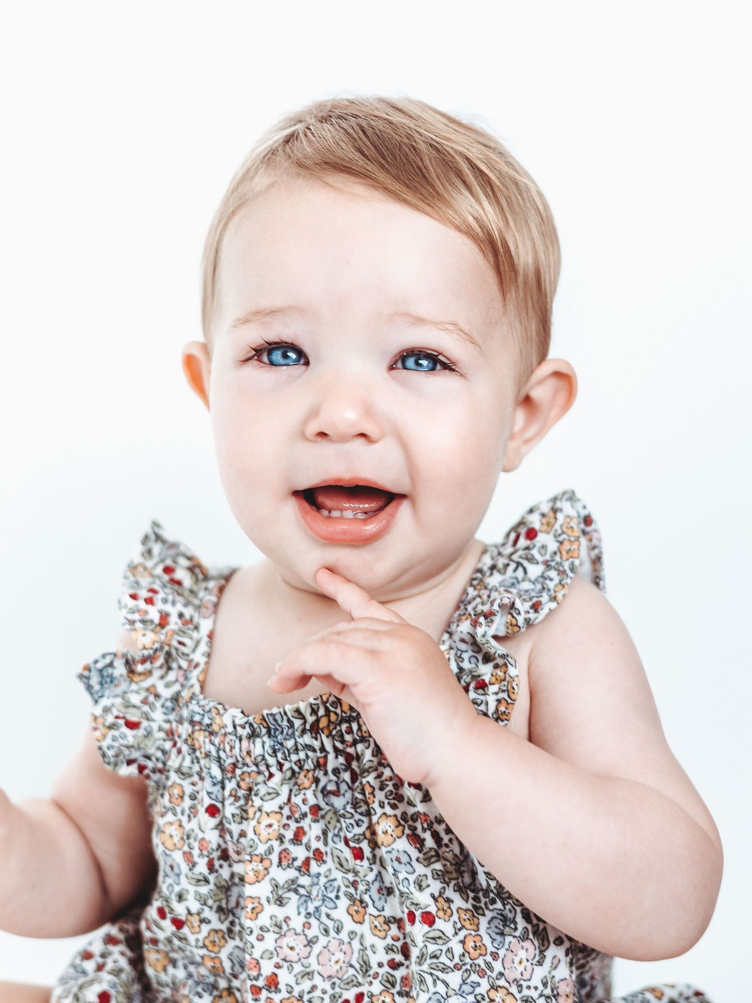 Adorable baby in floral dress smiles and laughs while sitting against white background in three progressive shots.