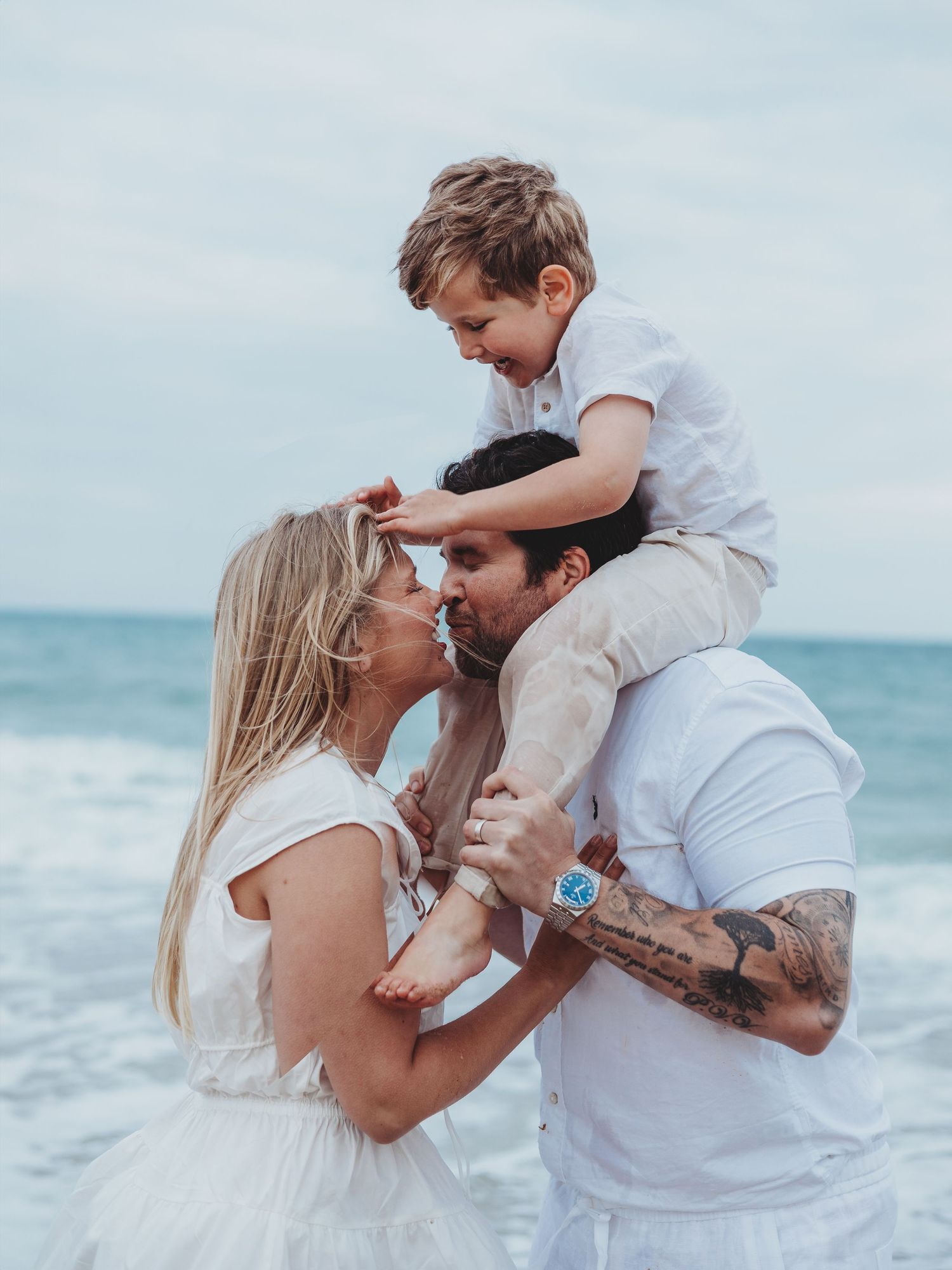 Family of three in white outfits at beach, parents sharing a kiss while young boy sits on dad's shoulders.