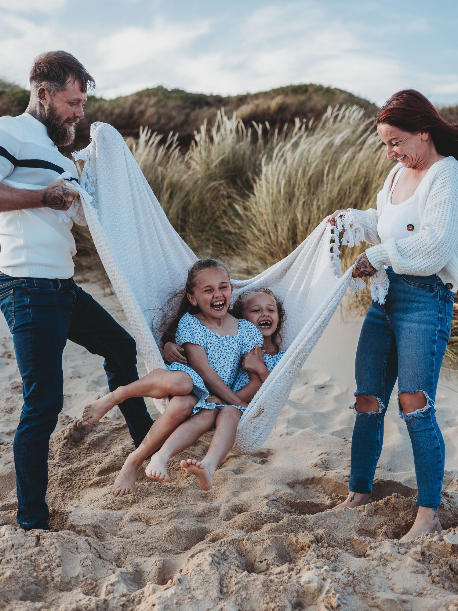 Family laughing on sandy beach as parents swing two girls in a white blanket hammock near grass dunes.