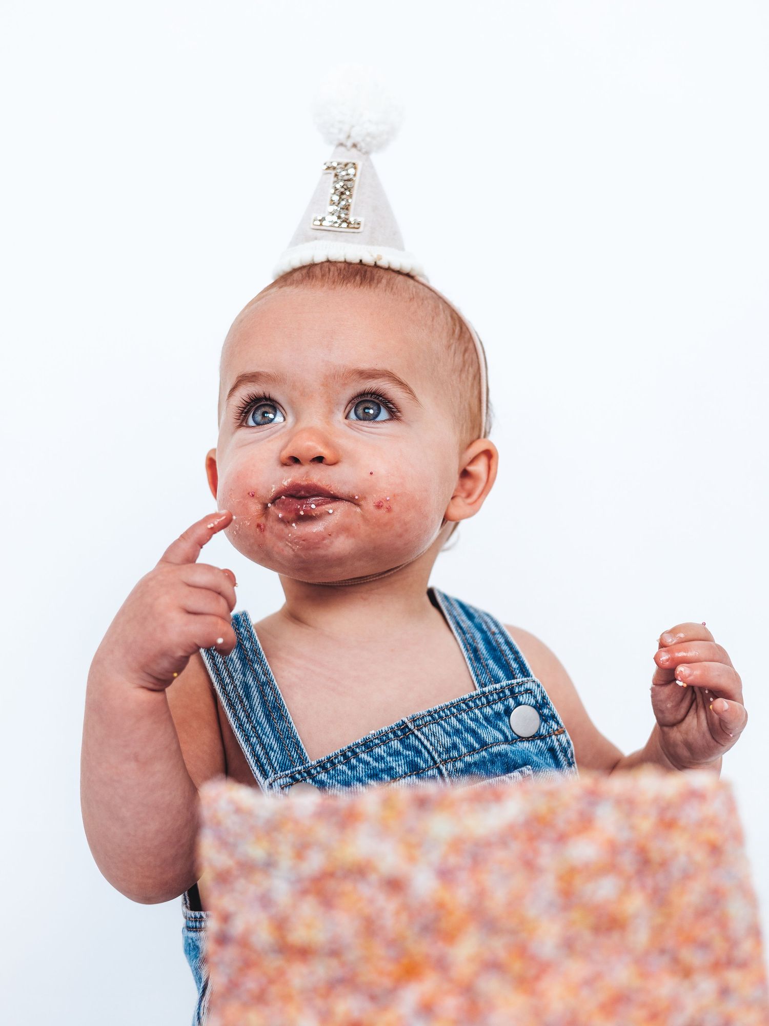 Baby in denim overalls and party hat eating birthday cake with chocolate frosting on face.