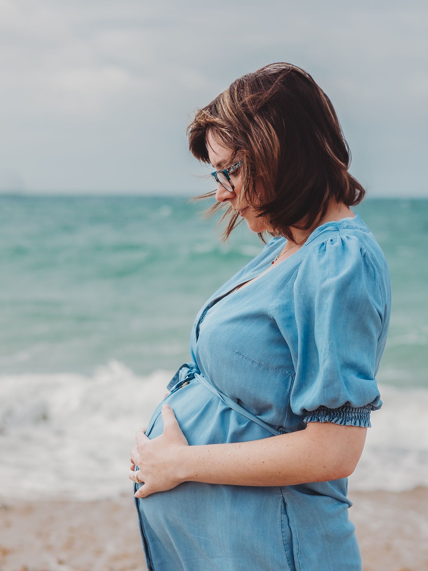A pregnant woman in a blue dress stands at the beach with turquoise waves and wind-swept hair.