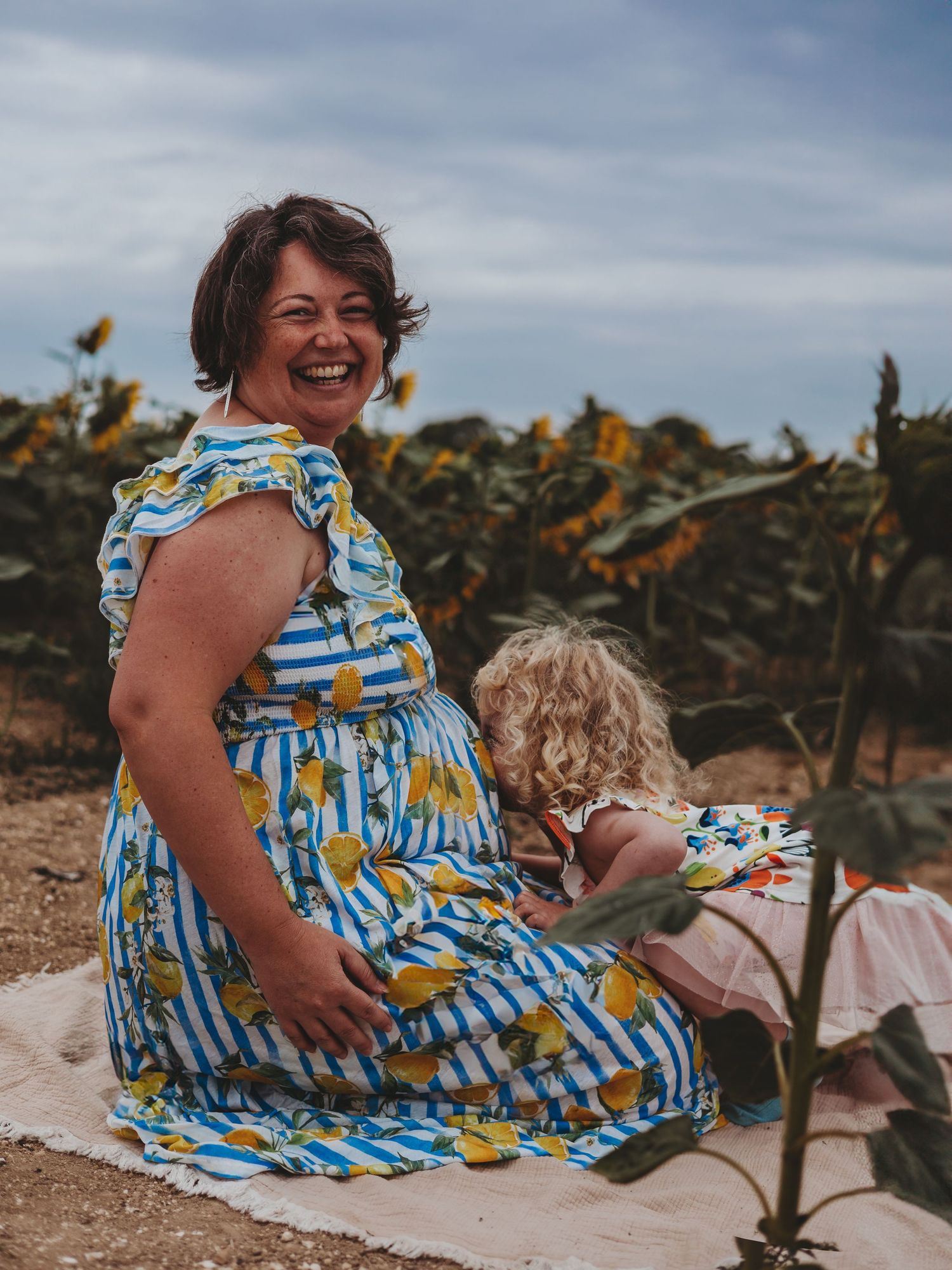 A person in a blue and yellow floral dress relaxes on a beach with sunflowers in the background during golden hour.