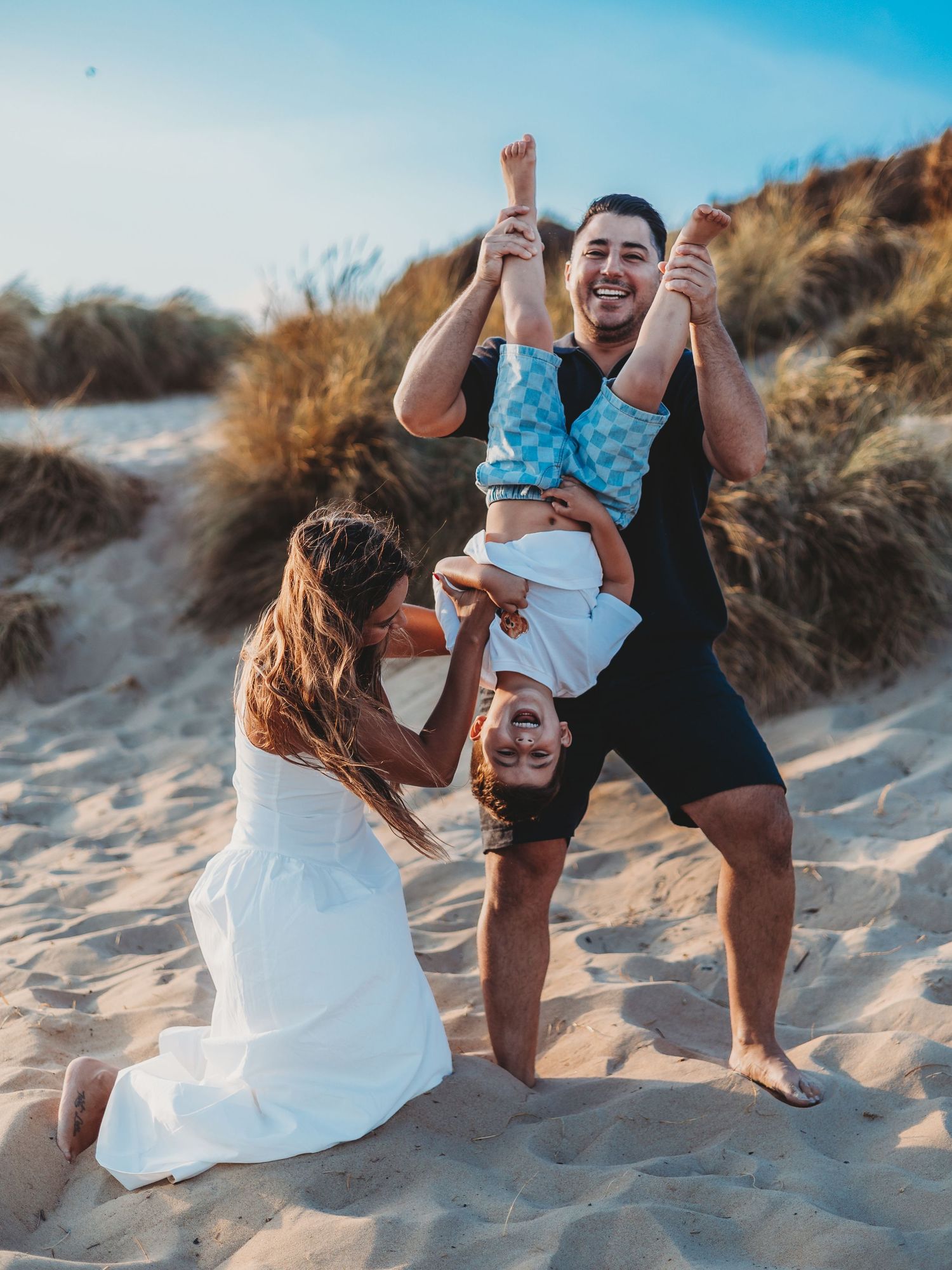 Family playing on sandy beach dunes, man holding child upside down while woman in white dress laughs joyfully.
