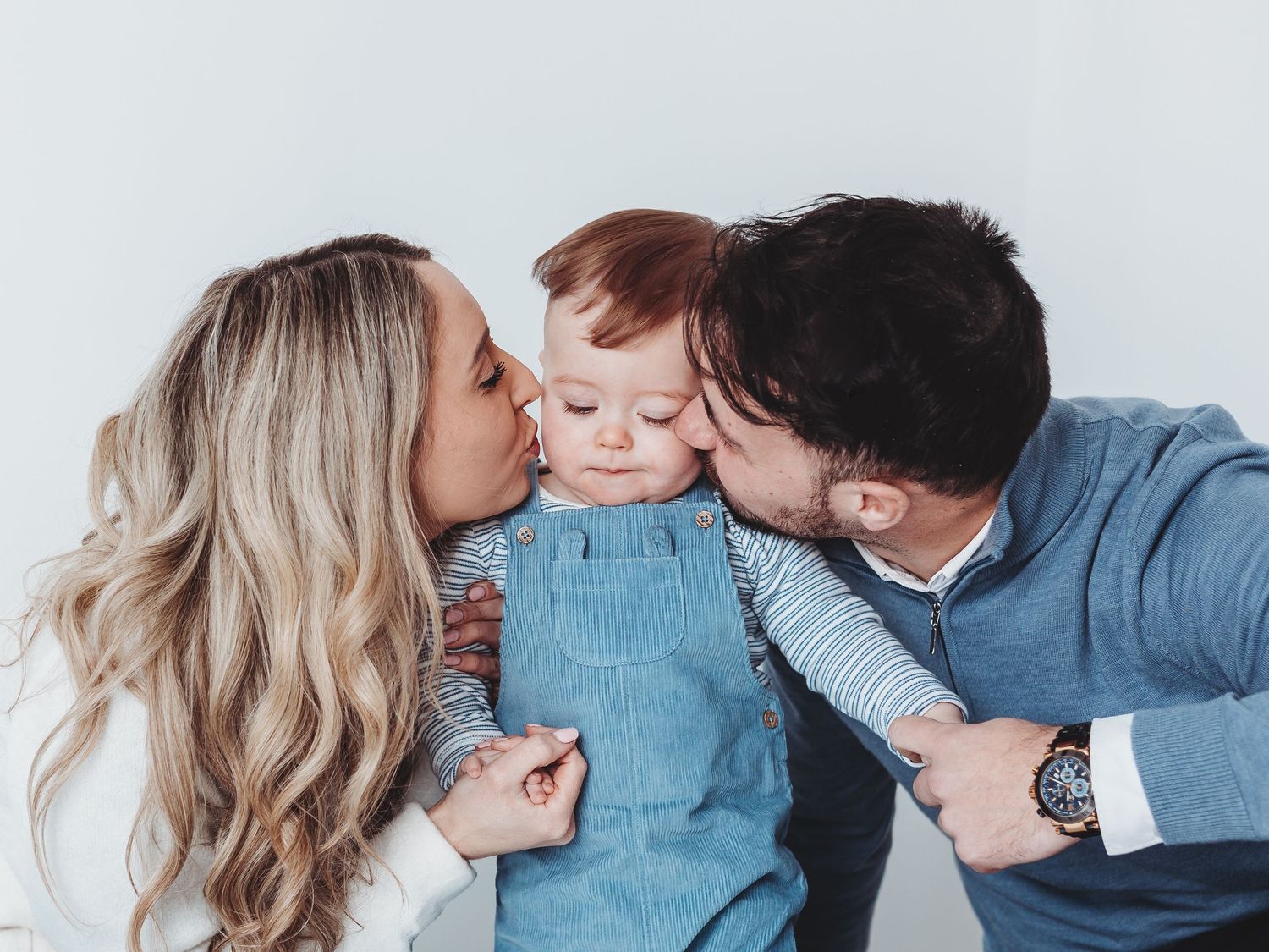 Parents kissing their adorable red-haired toddler on the cheeks in a bright studio family portrait.