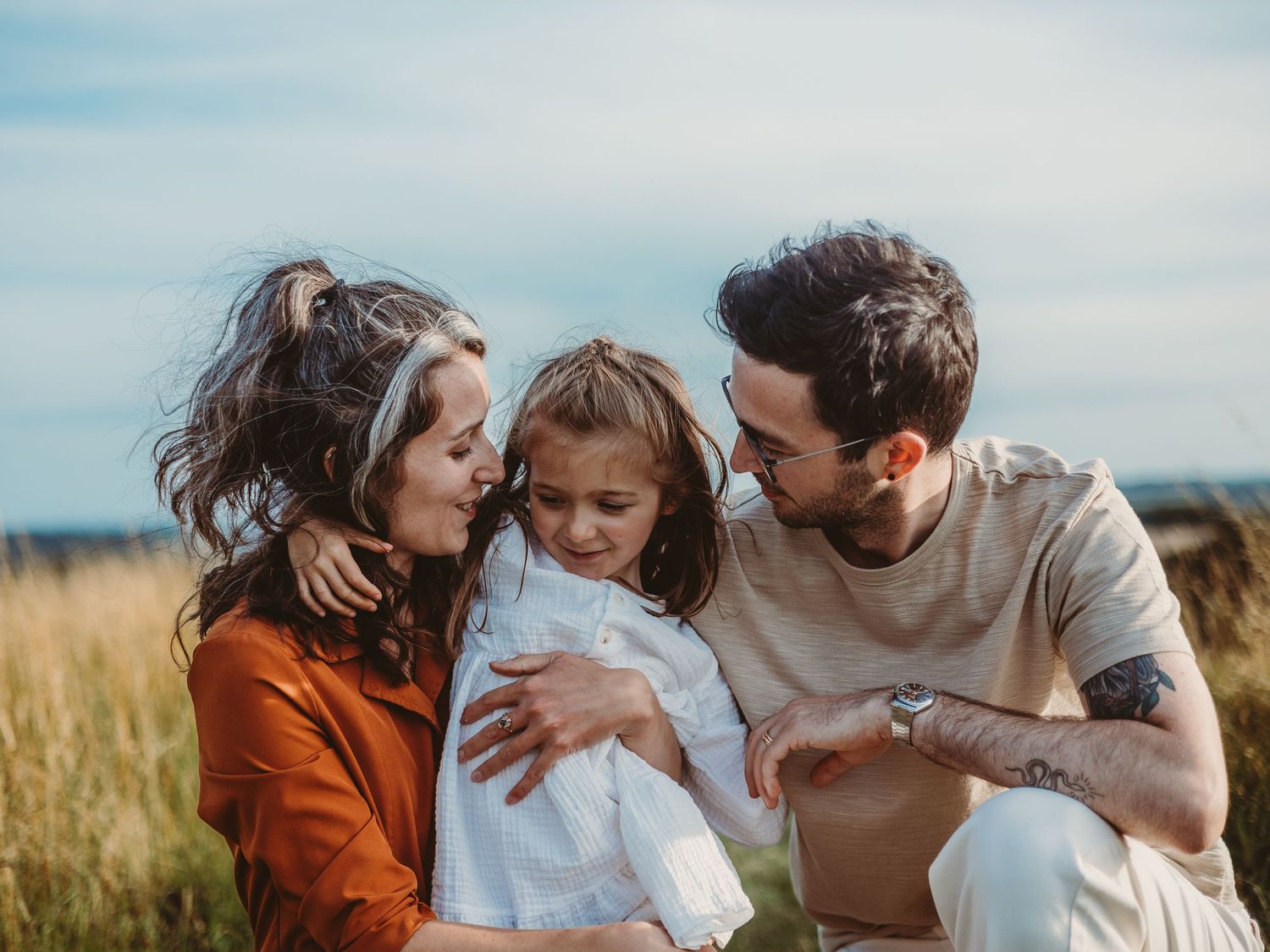 A loving family of three shares a tender moment in a golden field, parents embracing their young daughter outdoors.