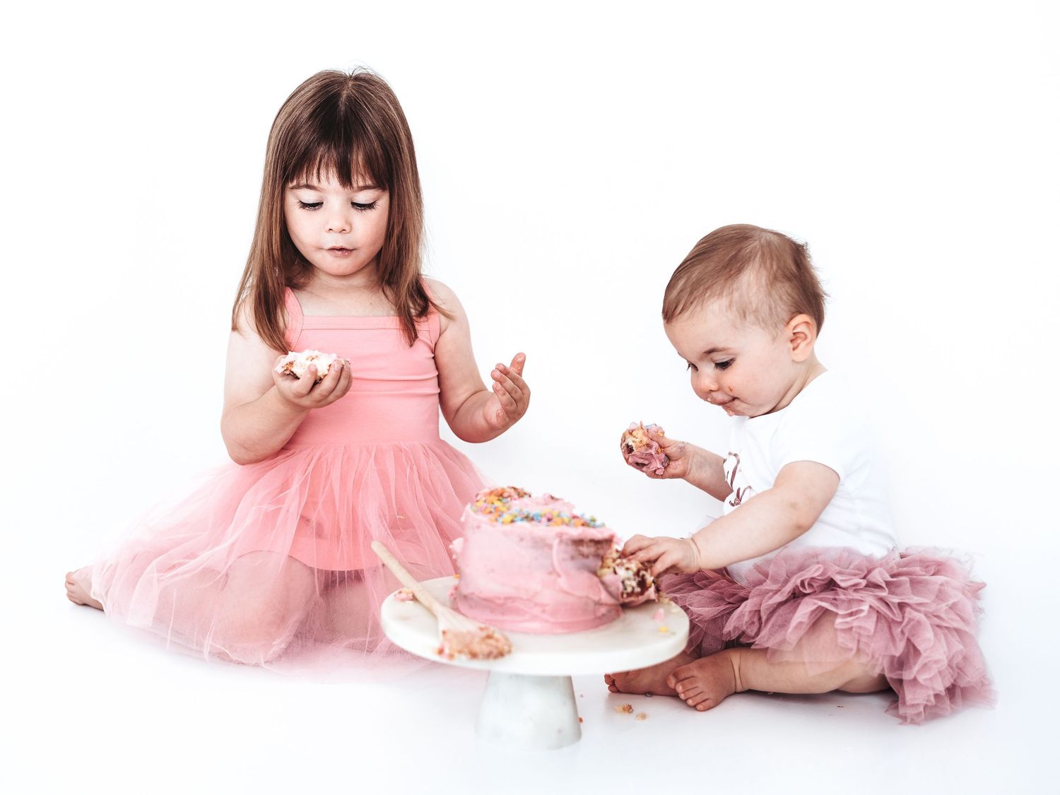 Two young girls in pink dresses smashing a pink birthday cake on a white background.