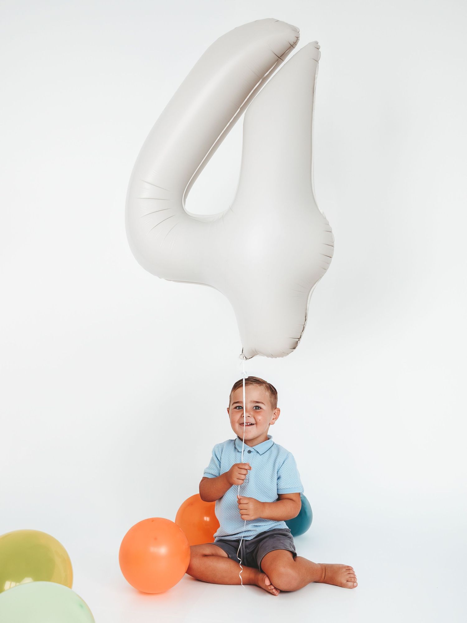 A toddler boy sits on a white floor holding a large number 4 balloon, surrounded by colorful balloons for his 4th birthday.