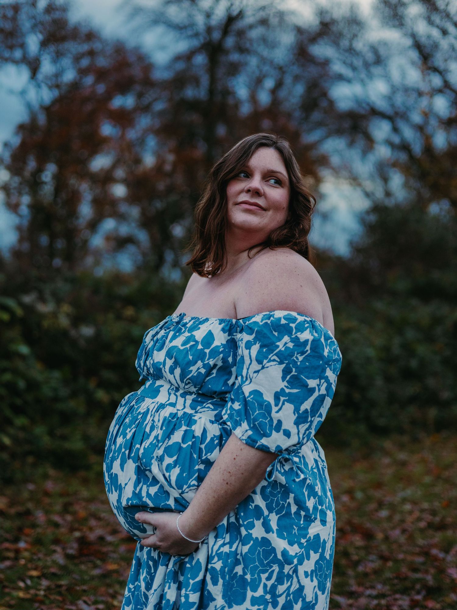 A person in a blue and white floral dress poses thoughtfully against an autumn forest backdrop during golden hour.