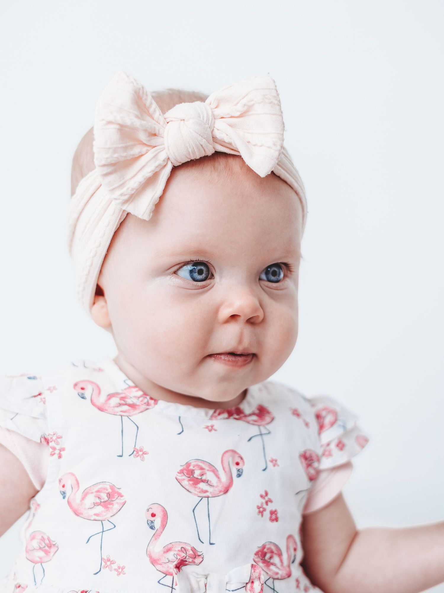 A baby wearing a pink bow headband and white floral dress smiles sweetly in a series of close-up photos.