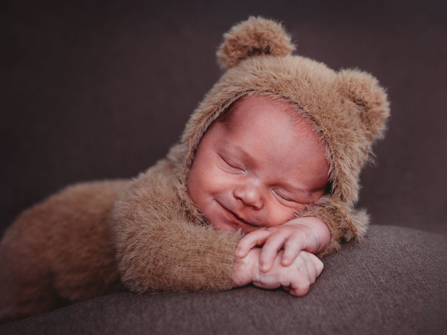 A sleeping newborn baby wearing a brown teddy bear hat rests peacefully on a dark gray blanket.