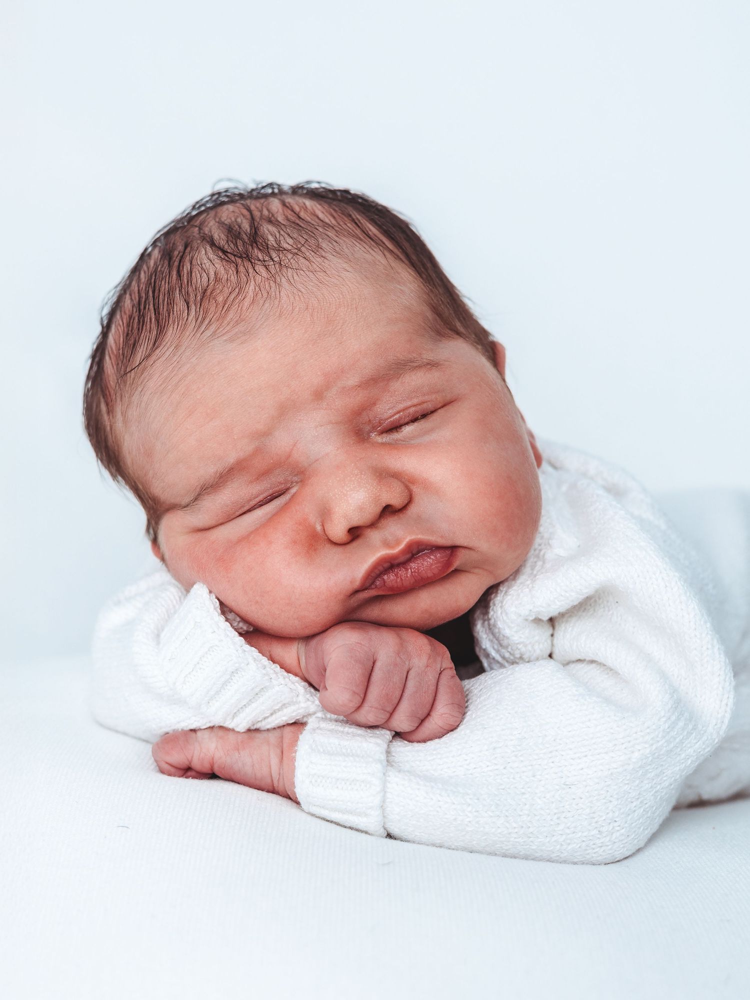 A sleeping newborn baby wearing a white sweater rests peacefully against a light blue background.