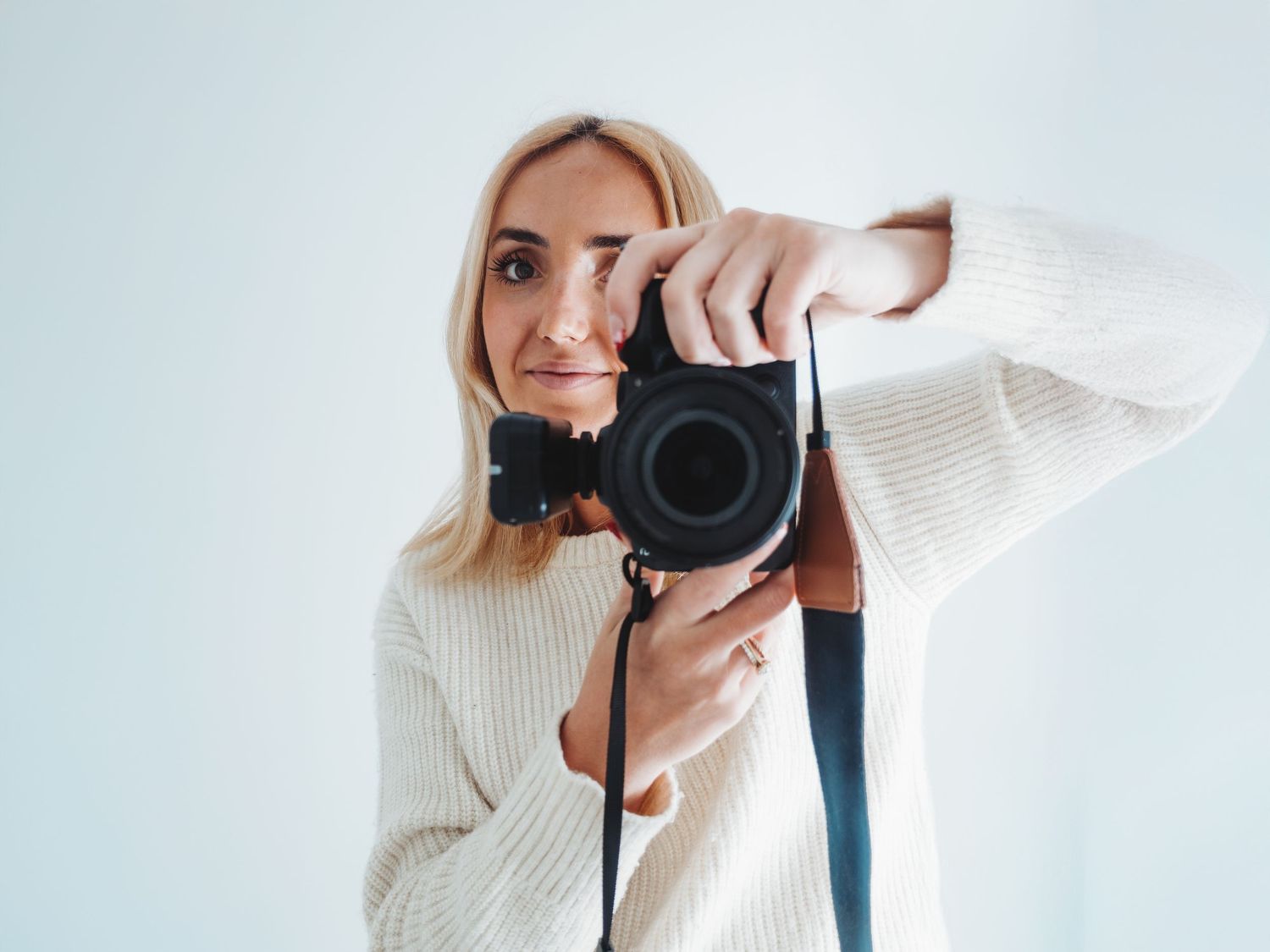 A photographer in a white sweater takes self-portraits with a professional camera against a bright background.