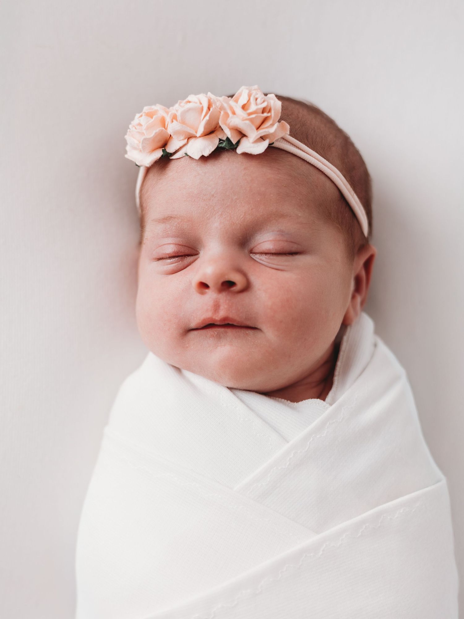 Sleeping newborn baby wrapped in white swaddle blanket wearing delicate pink floral headband.
