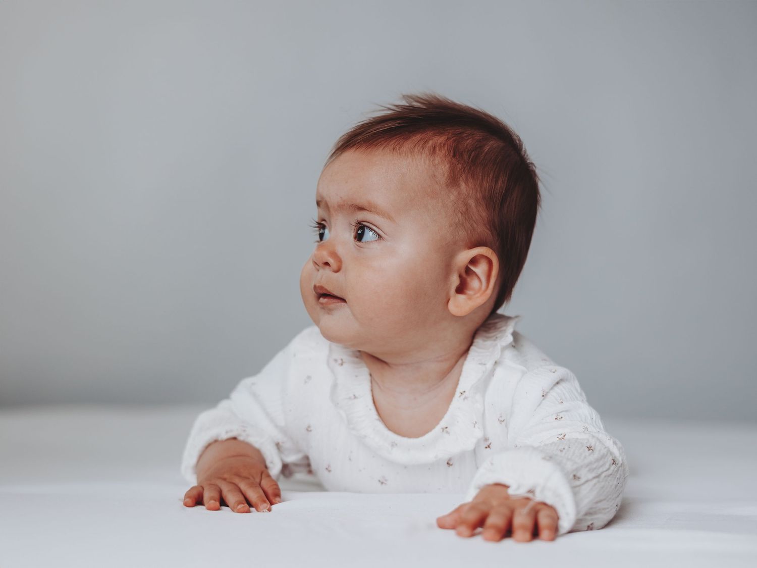 Adorable baby in white onesie doing tummy time, looking up curiously against a soft gray background.