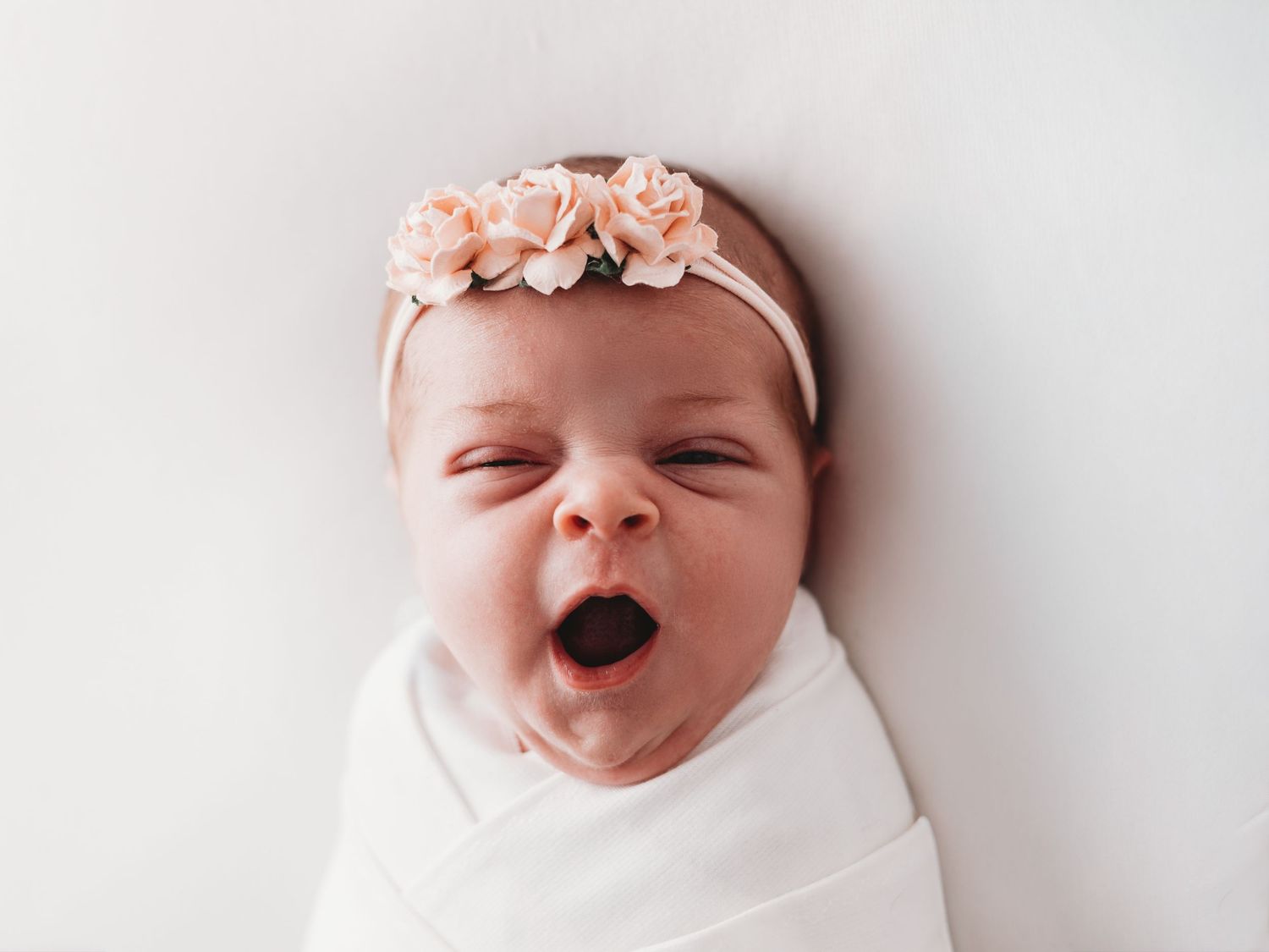 A newborn baby wrapped in a white blanket yawns while wearing a delicate pink floral headband.
