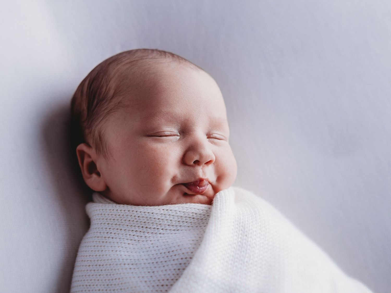 Peaceful sleeping newborn baby wrapped snugly in soft white blanket against light background.