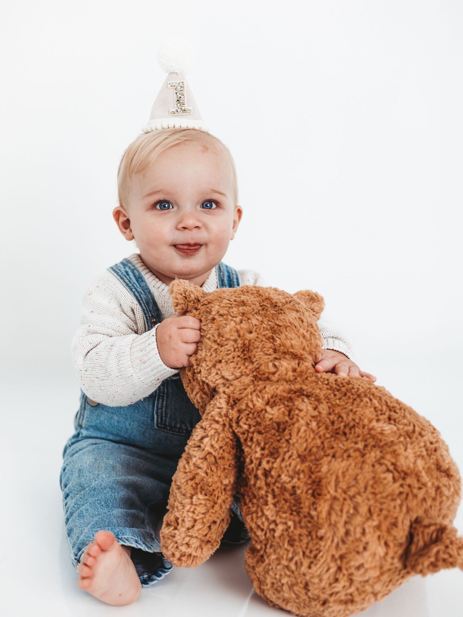 A smiling baby wearing a party hat holds a brown teddy bear, sitting against a white background.
