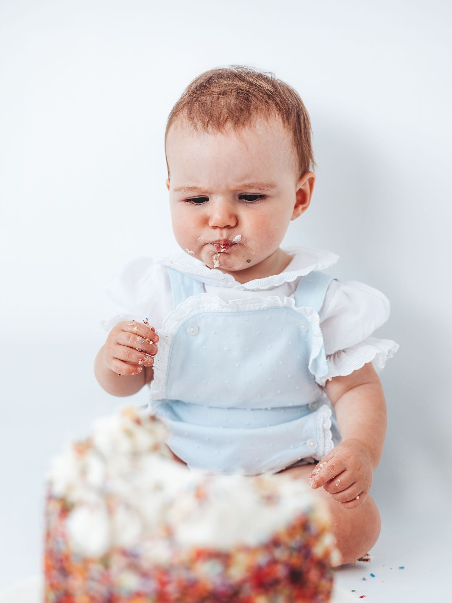 Baby in blue romper eating first birthday cake with white frosting and colorful sprinkles against white background.
