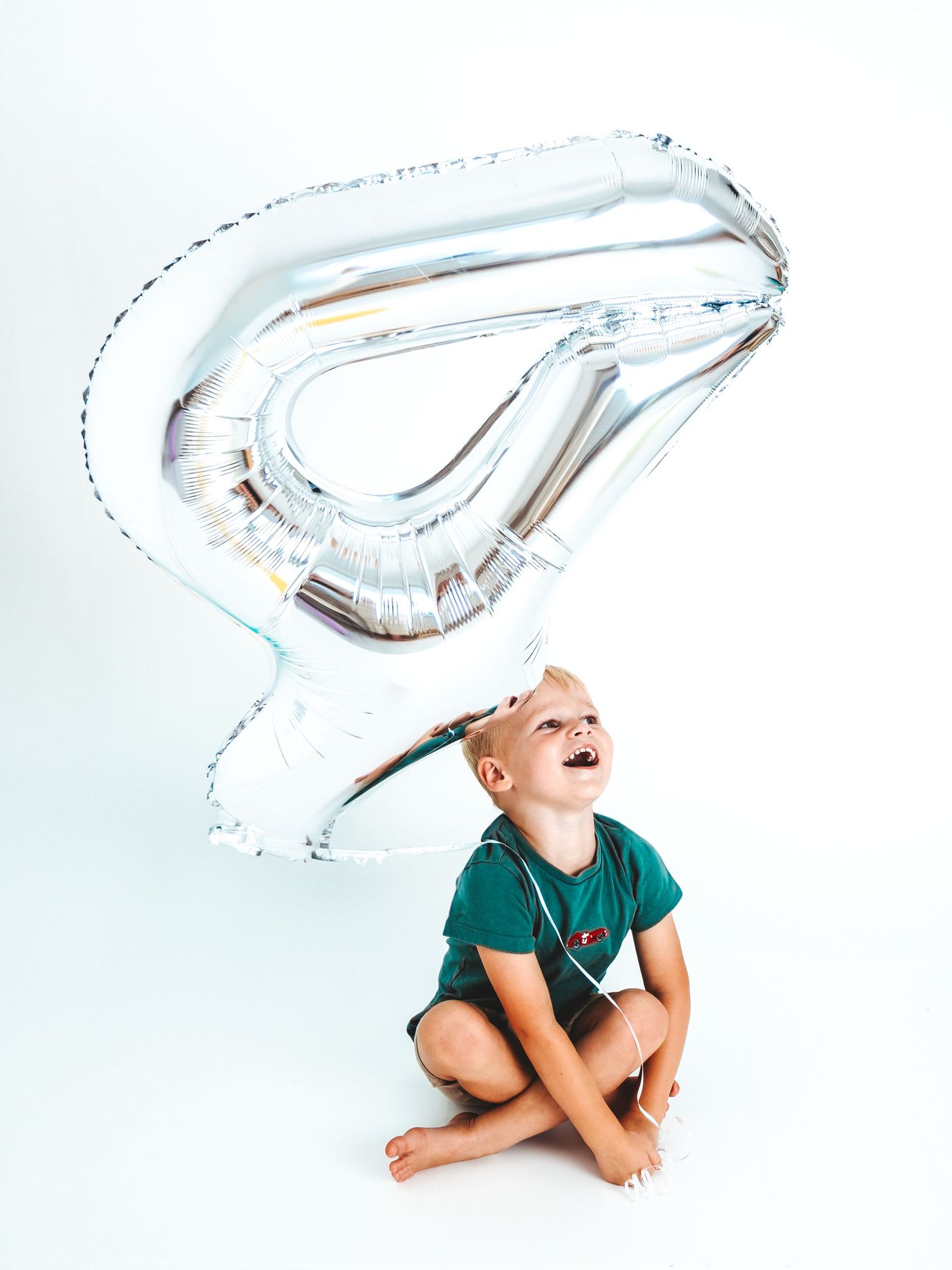 A laughing young boy sits cross-legged under a large silver number 4 balloon on a white background.