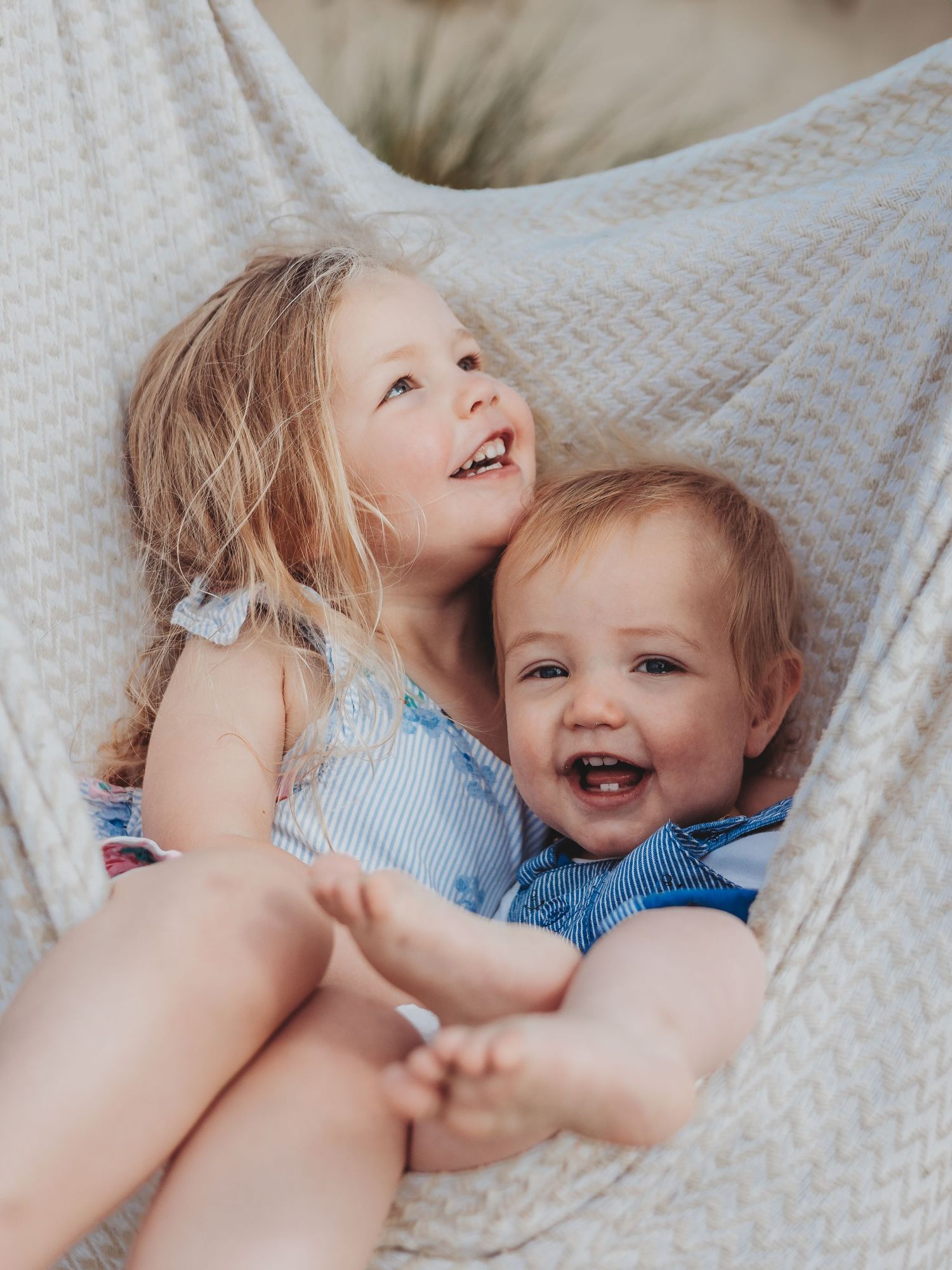 Two happy young children laughing together in a cozy hammock, wearing blue outfits on a warm sunny day.