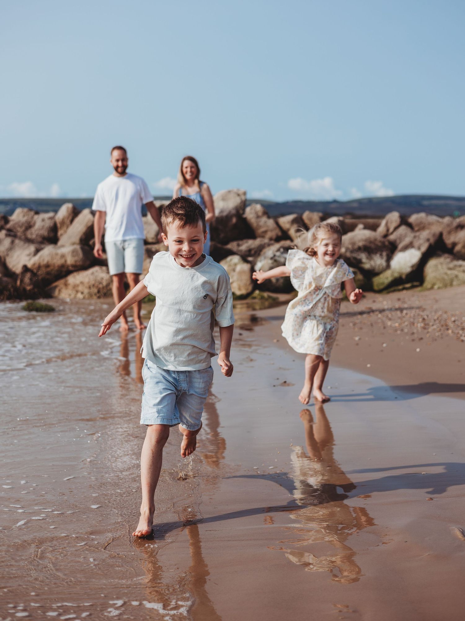 Family of four enjoying a sunny beach day, with two young children running along the shoreline near rocky shores.