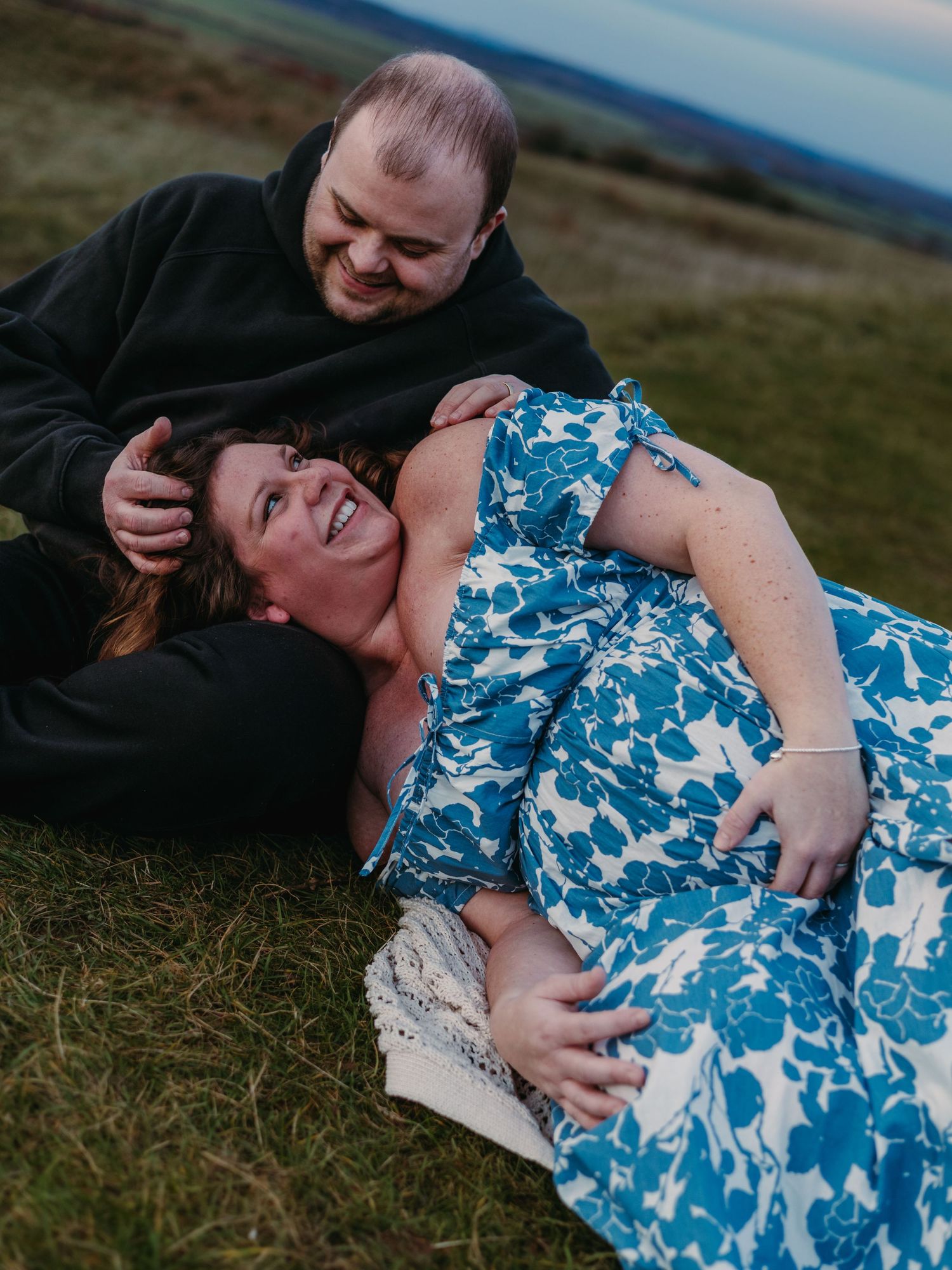 A couple shares intimate moments in a grassy field while wearing a blue and white floral dress and dark clothing.