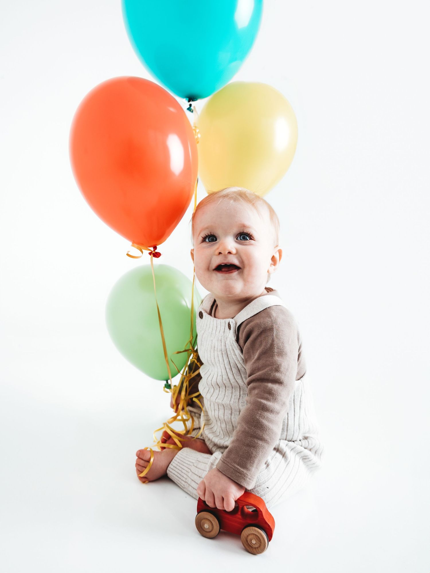 Happy baby sitting with colorful balloons and a red toy car, smiling joyfully in a bright white studio setting.