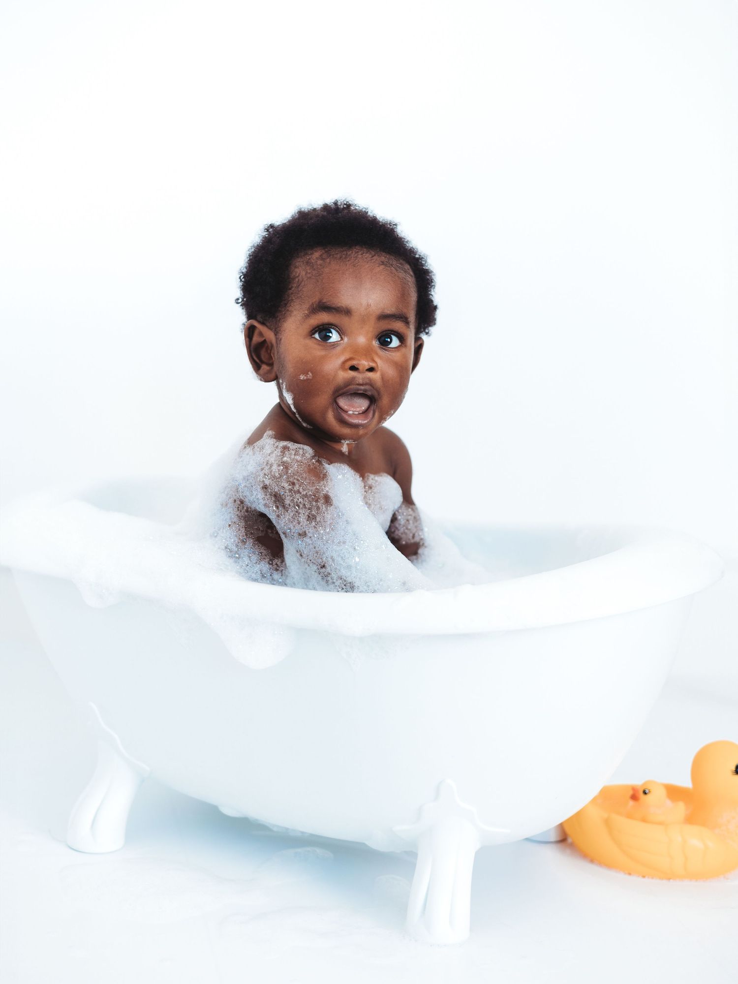 Adorable Black baby sitting in a small white clawfoot bathtub with bubbles, mouth open in surprise, rubber duck nearby.