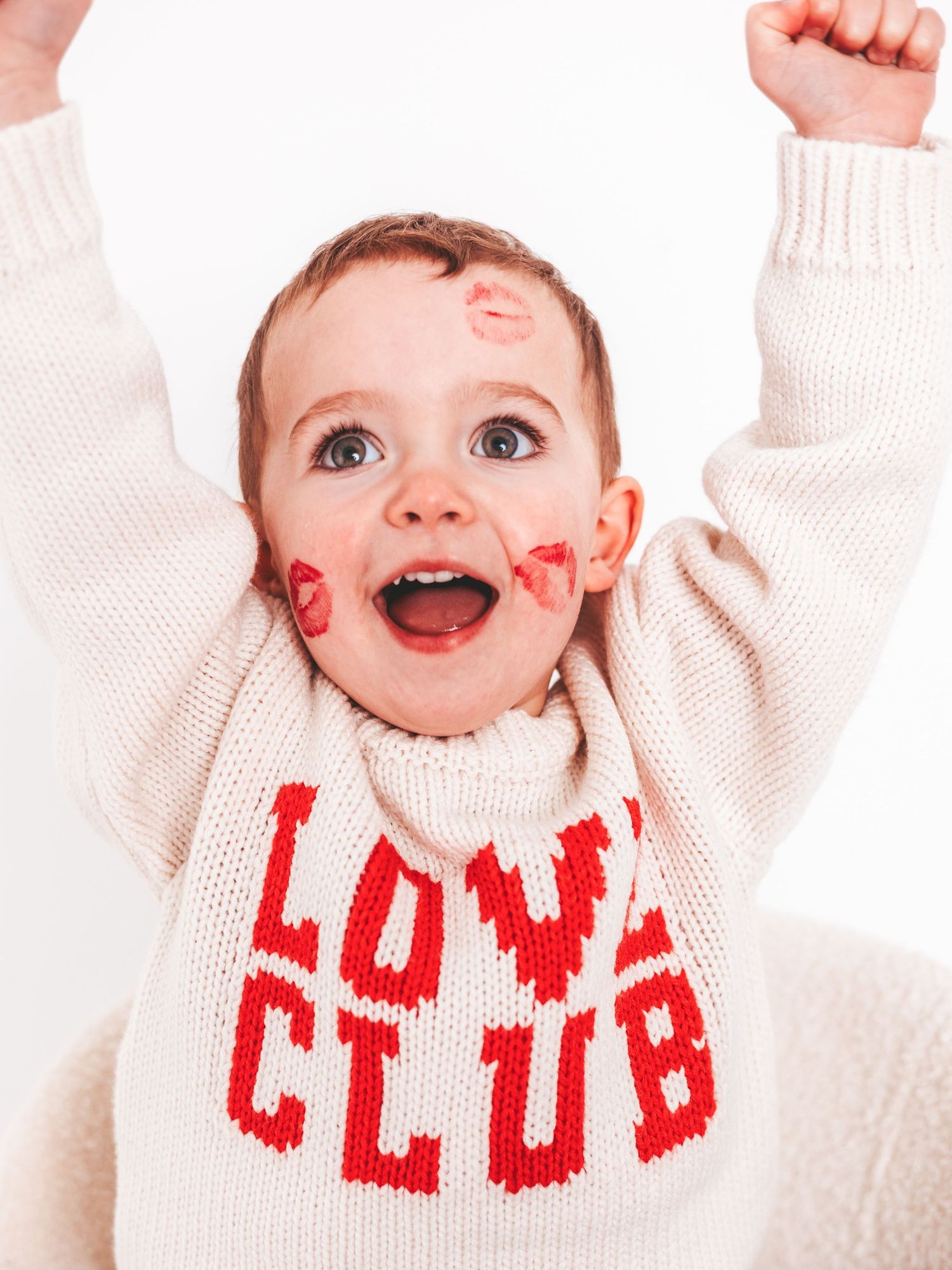 Joyful baby with lipstick kiss marks on face wearing a cream Love Club sweater, arms raised in celebration.