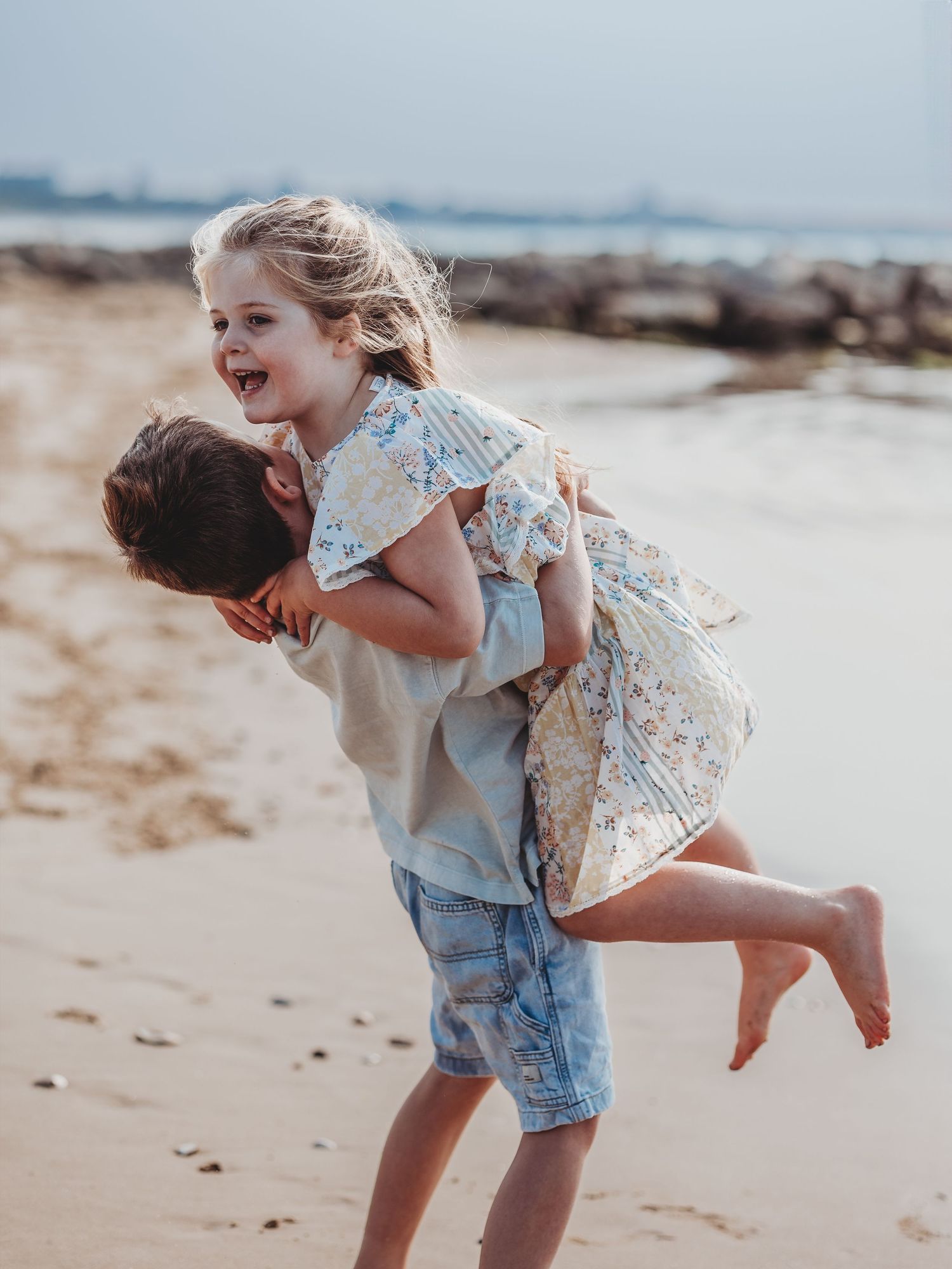 Two children laughing and playing piggyback on a sandy beach, girl in floral dress giggling with joy.