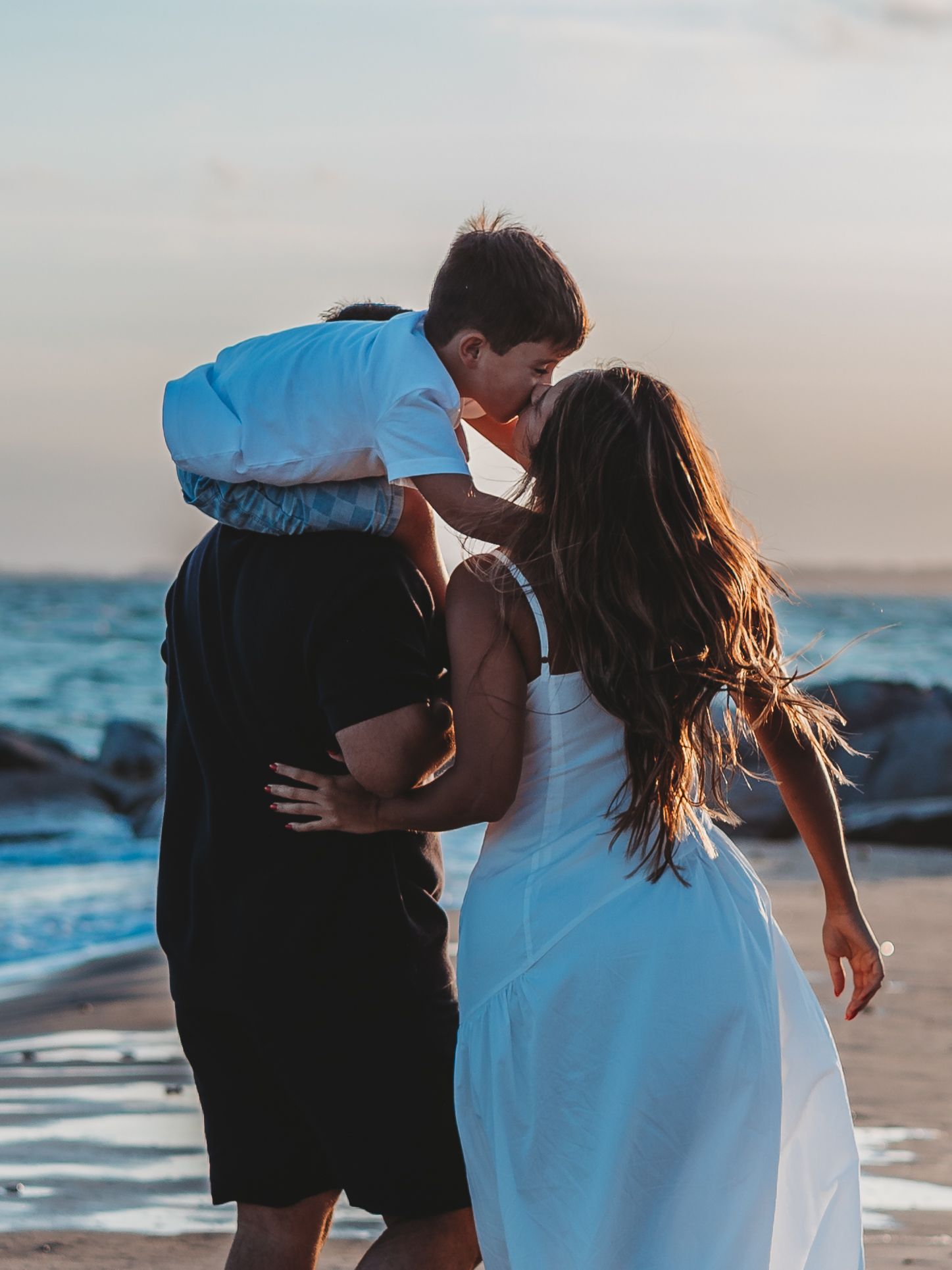 A couple shares a romantic kiss on the beach at sunset, woman in white dress, man in white shirt and black pants.