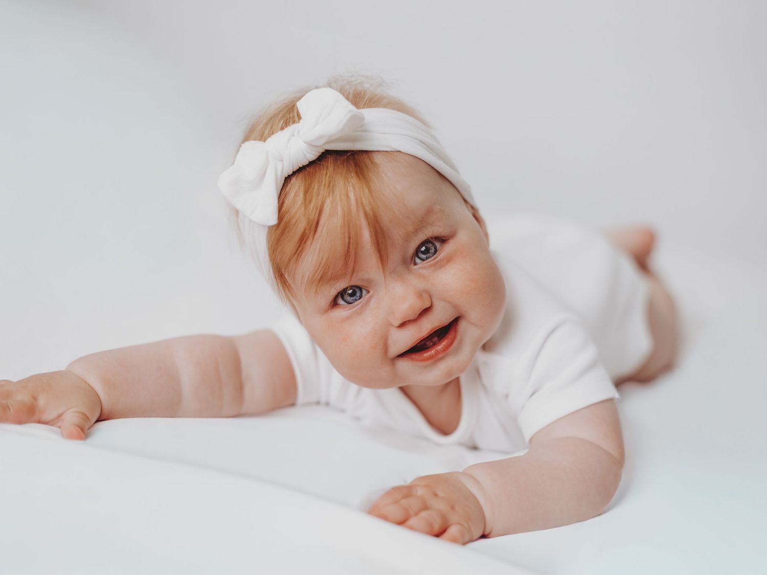 A baby in white clothing smiles while lying on soft white bedding.