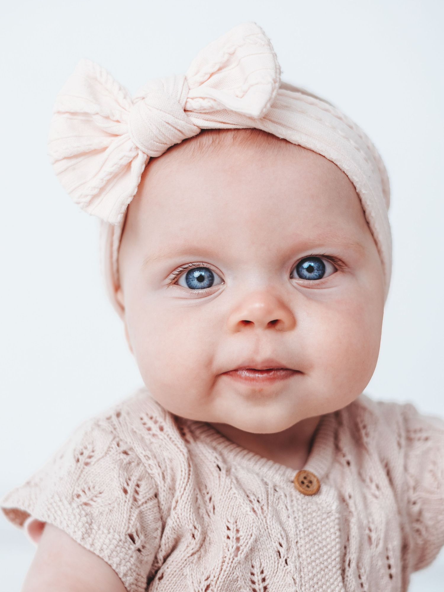 A baby wearing a light pink knit outfit and bow headband smiles sweetly at the camera.