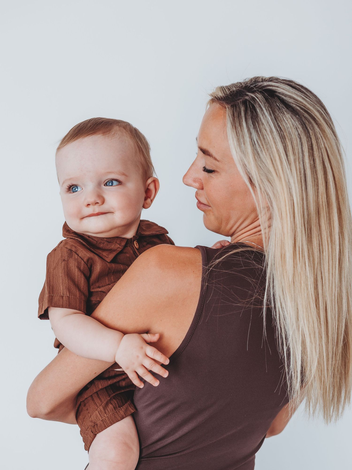 Blonde woman in brown dress lovingly holds a smiling baby in a brown shirt against a white background.