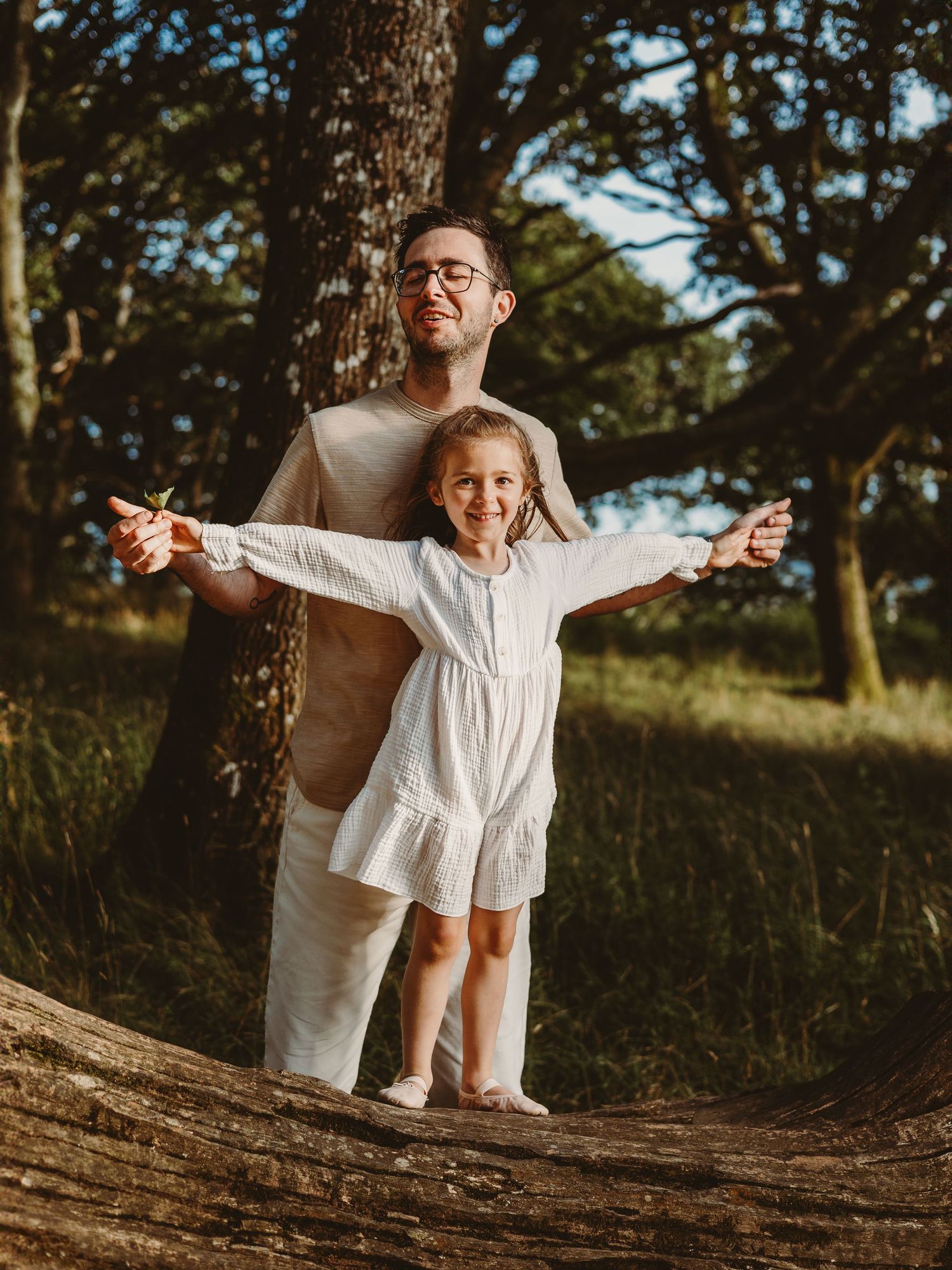 A father and young daughter in white outfits stand on a log in a sunlit forest, arms outstretched joyfully.