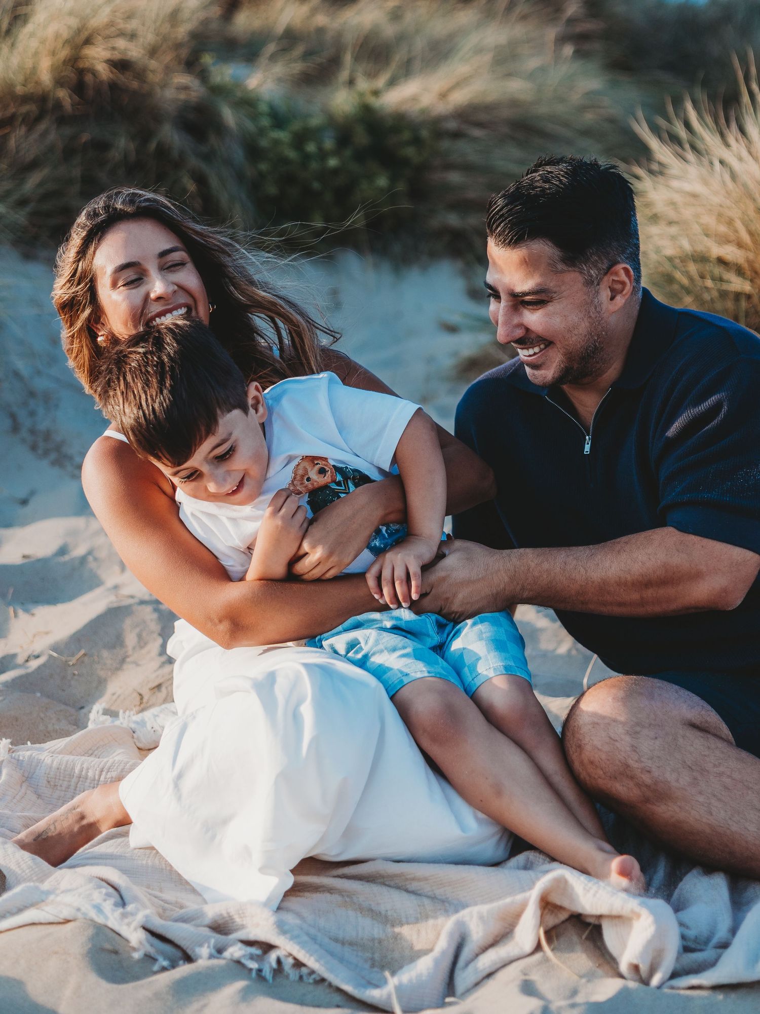 Happy family of three laughing and cuddling on a sandy beach blanket surrounded by dunes at golden hour.