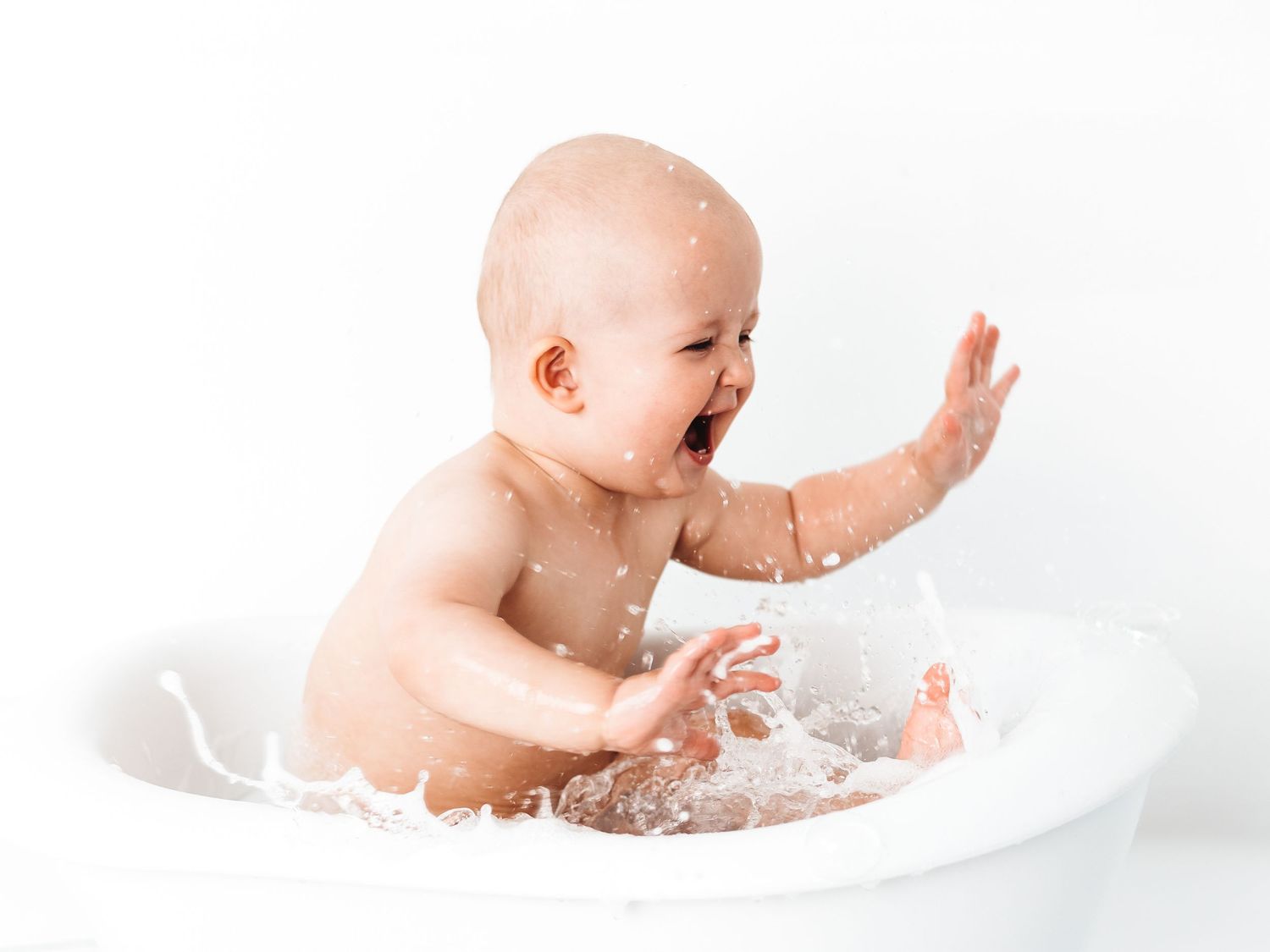 Laughing baby splashing water in a white bathtub during bath time.