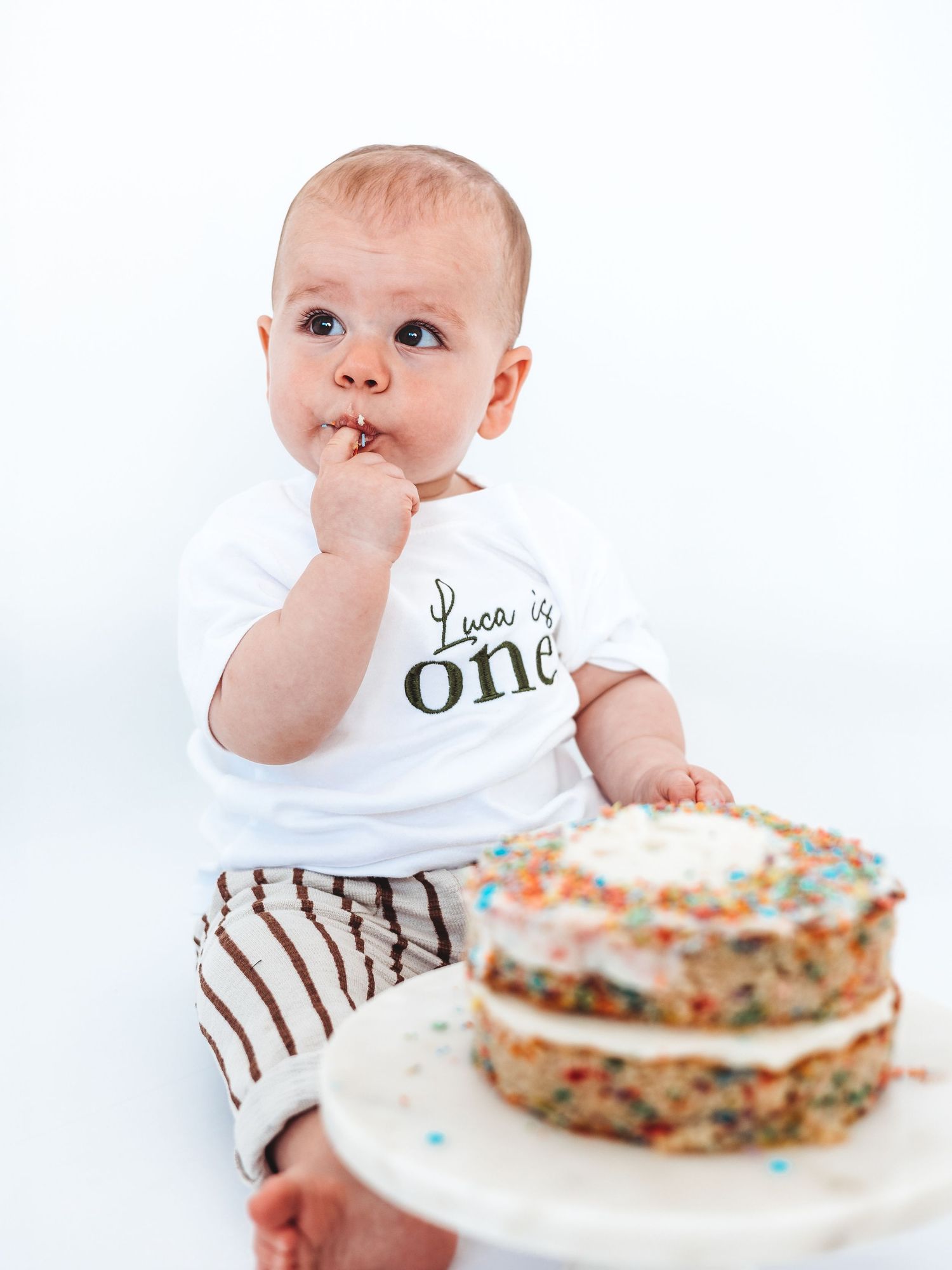 Baby Luca celebrates turning one in a white onesie beside a colorful sprinkle cake, finger in mouth.