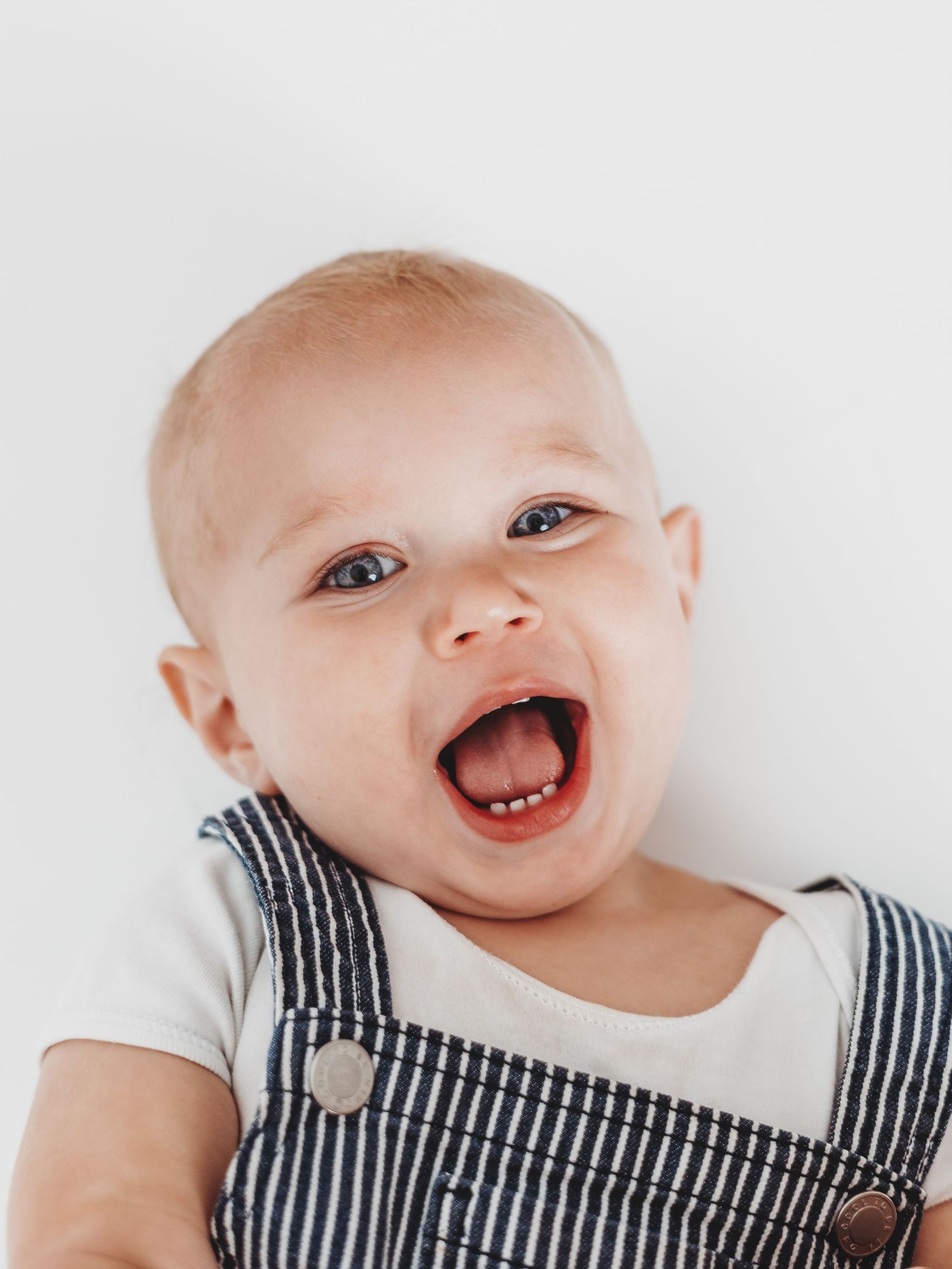 Happy baby wearing striped overalls laughs with pure joy against white background.
