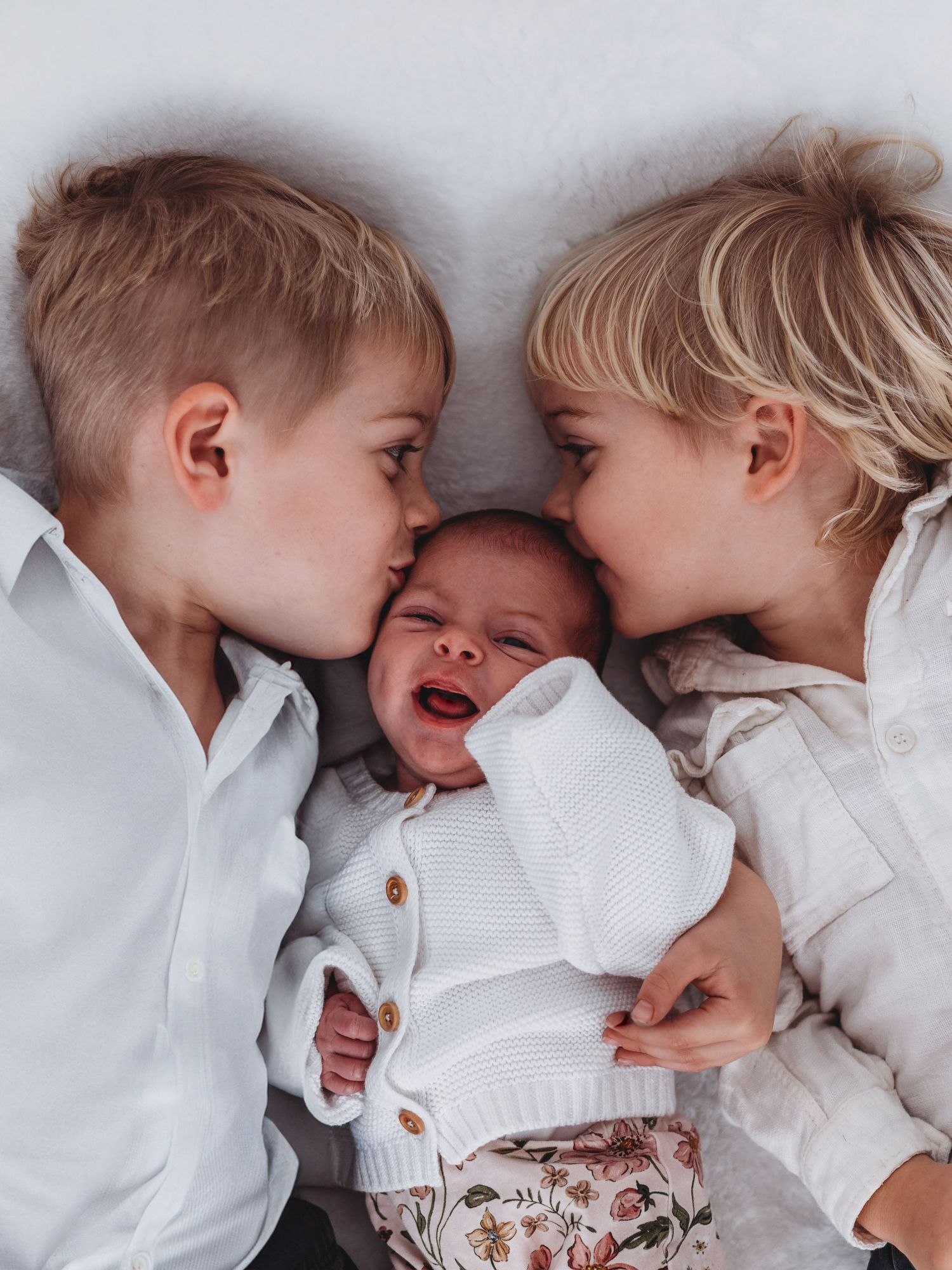 Two siblings in white shirts share sweet moments of affection while lying on light colored bedding.