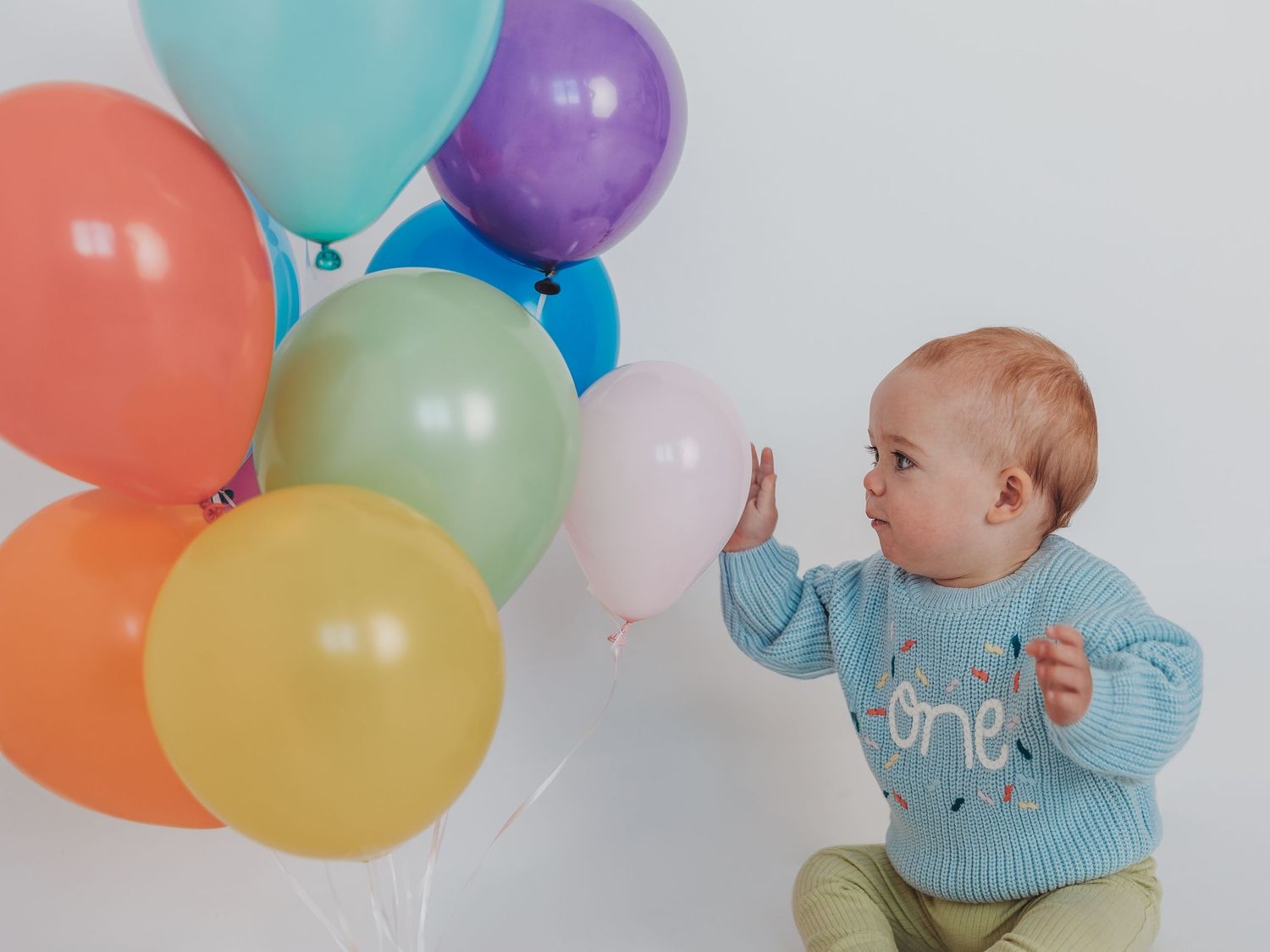Baby in light blue sweater plays with colorful balloons during a first birthday photo session.